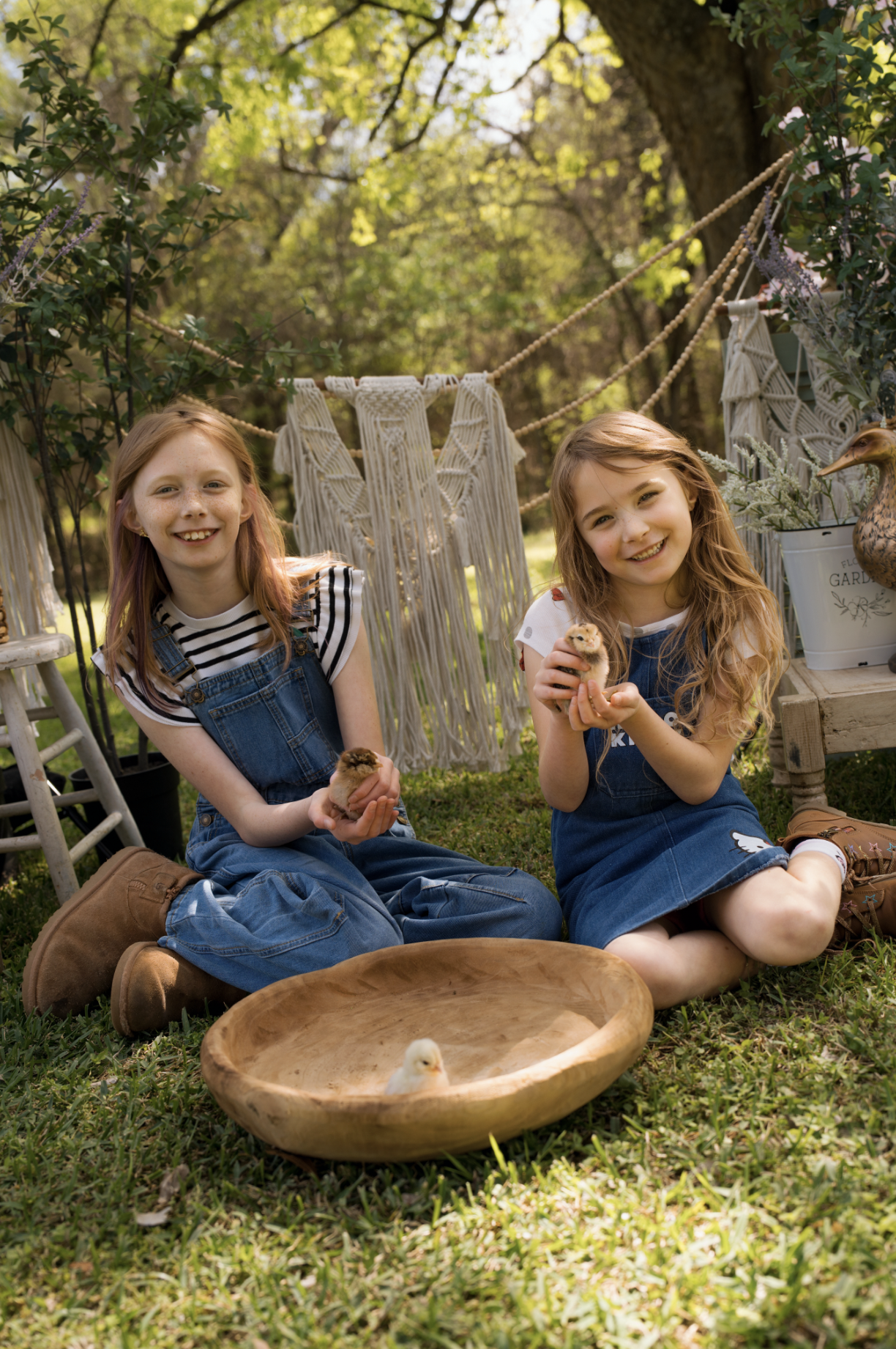 Two young girls sitting on grass outdoors, holding small chickens, with a wooden bowl containing a chick in front of them. They are smiling, surrounded by greenery and garden decor, including a macrame backdrop and plant pots.