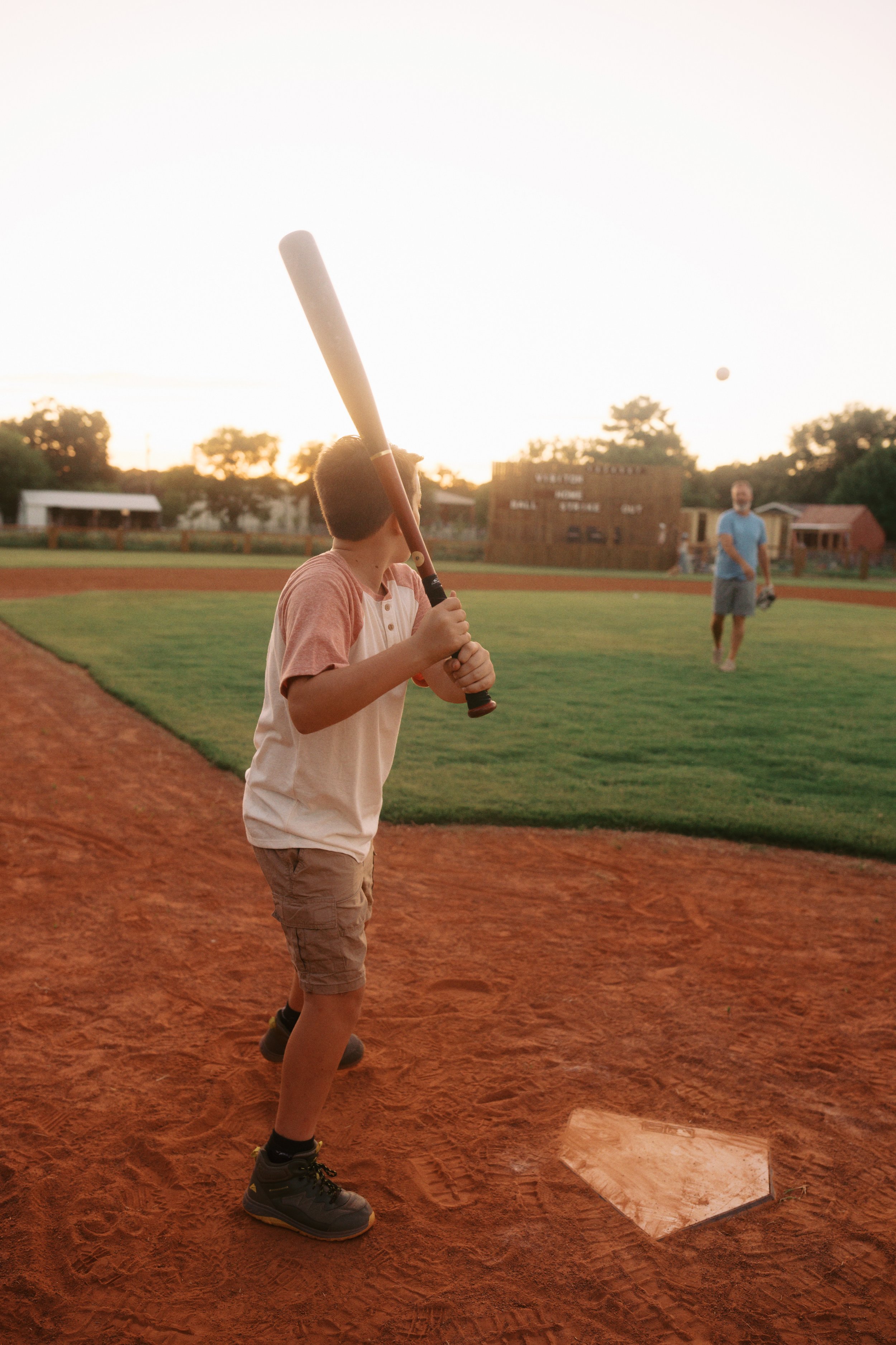 A young boy in a baseball uniform holding a bat behind his head, preparing to swing, on a baseball field during sunset. An adult man is in the background, standing on the grass.