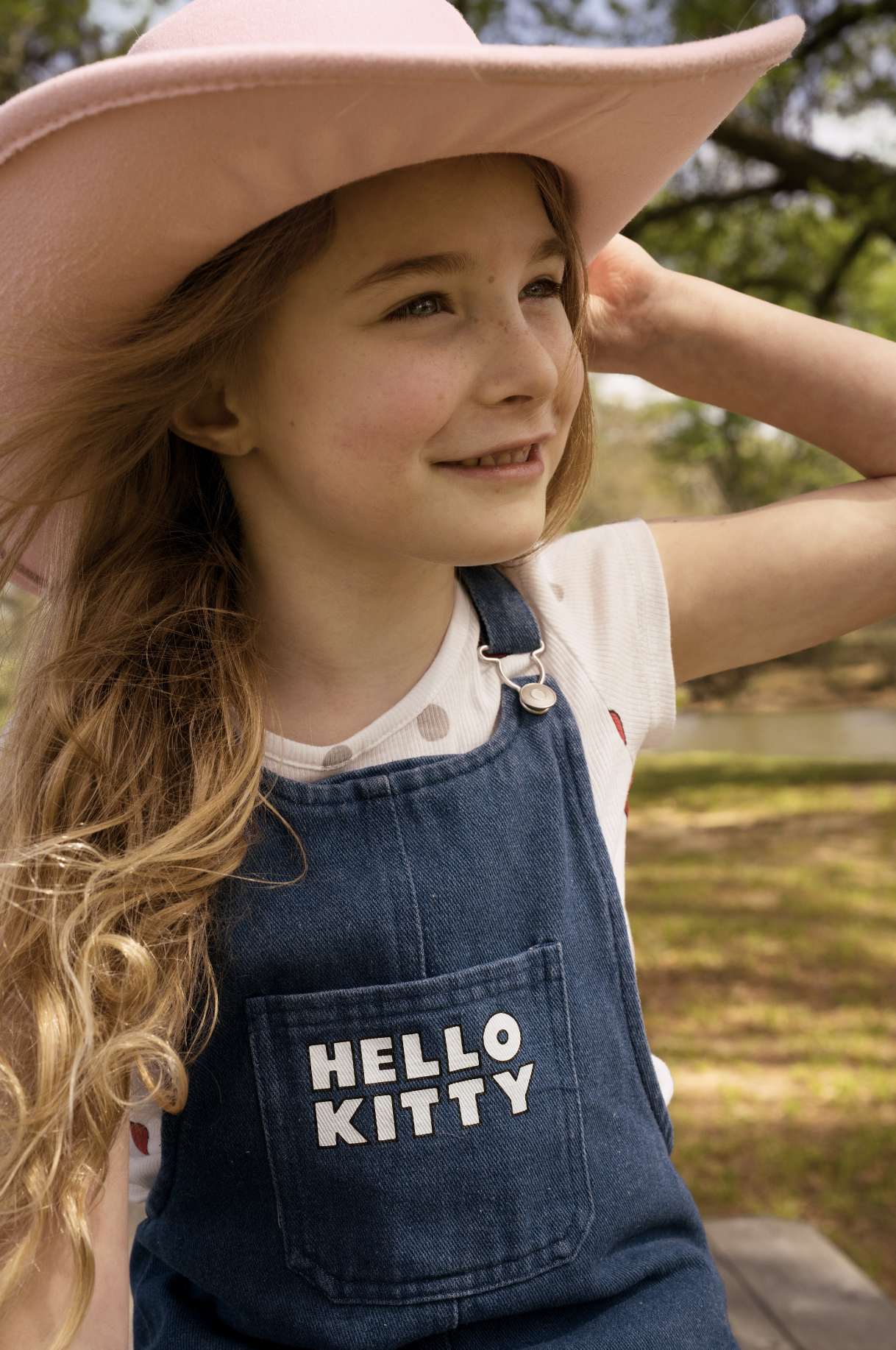 Young girl with long curly hair, wearing a pink wide-brimmed hat, white shirt with polka dots, and a blue Hello Kitty bib, standing outdoors with trees and a park in the background.