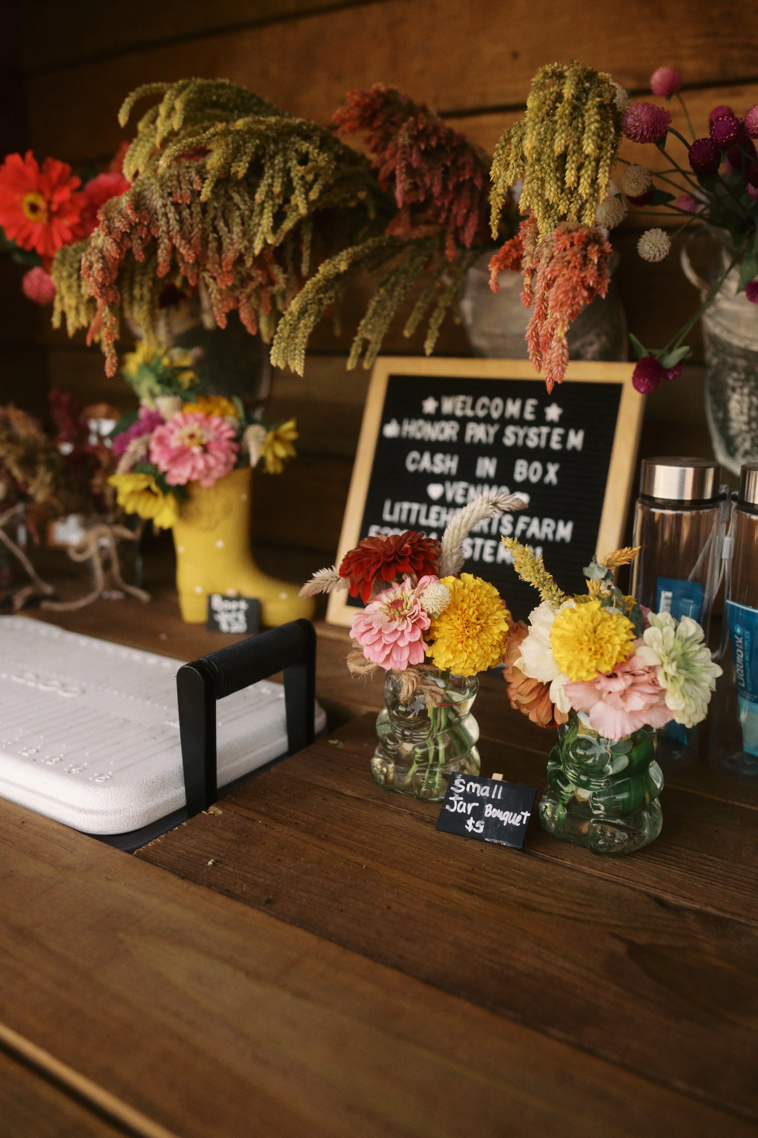 Various colorful flowers in glass jars on a wooden table, with a small chalkboard sign that reads 'Small Jar Bouquet $5'.