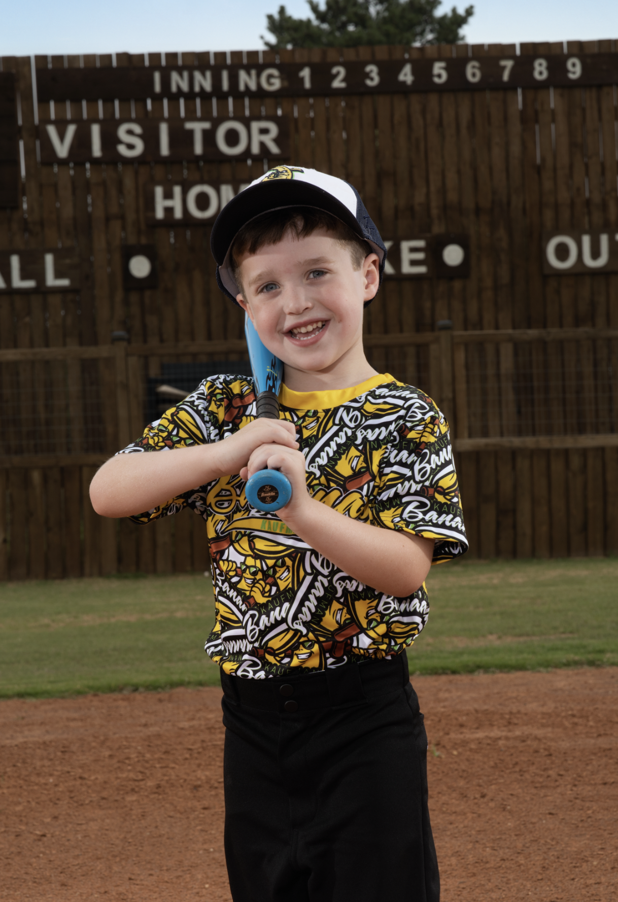 A young boy smiling at a baseball field holding a blue baseball bat, wearing a colorful baseball jersey, black pants, and a black and white cap.