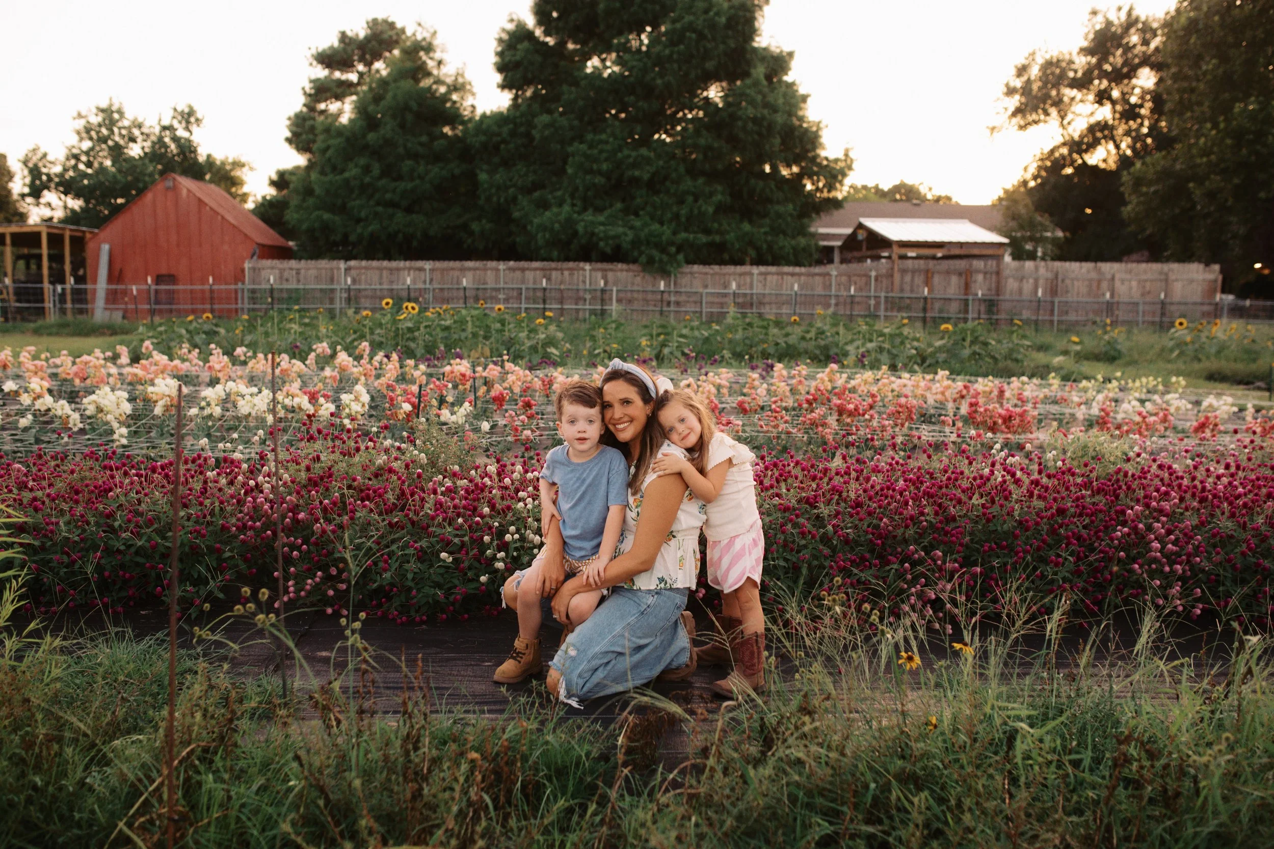 A woman with two young children in a flower garden during sunset, smiling and hugging.