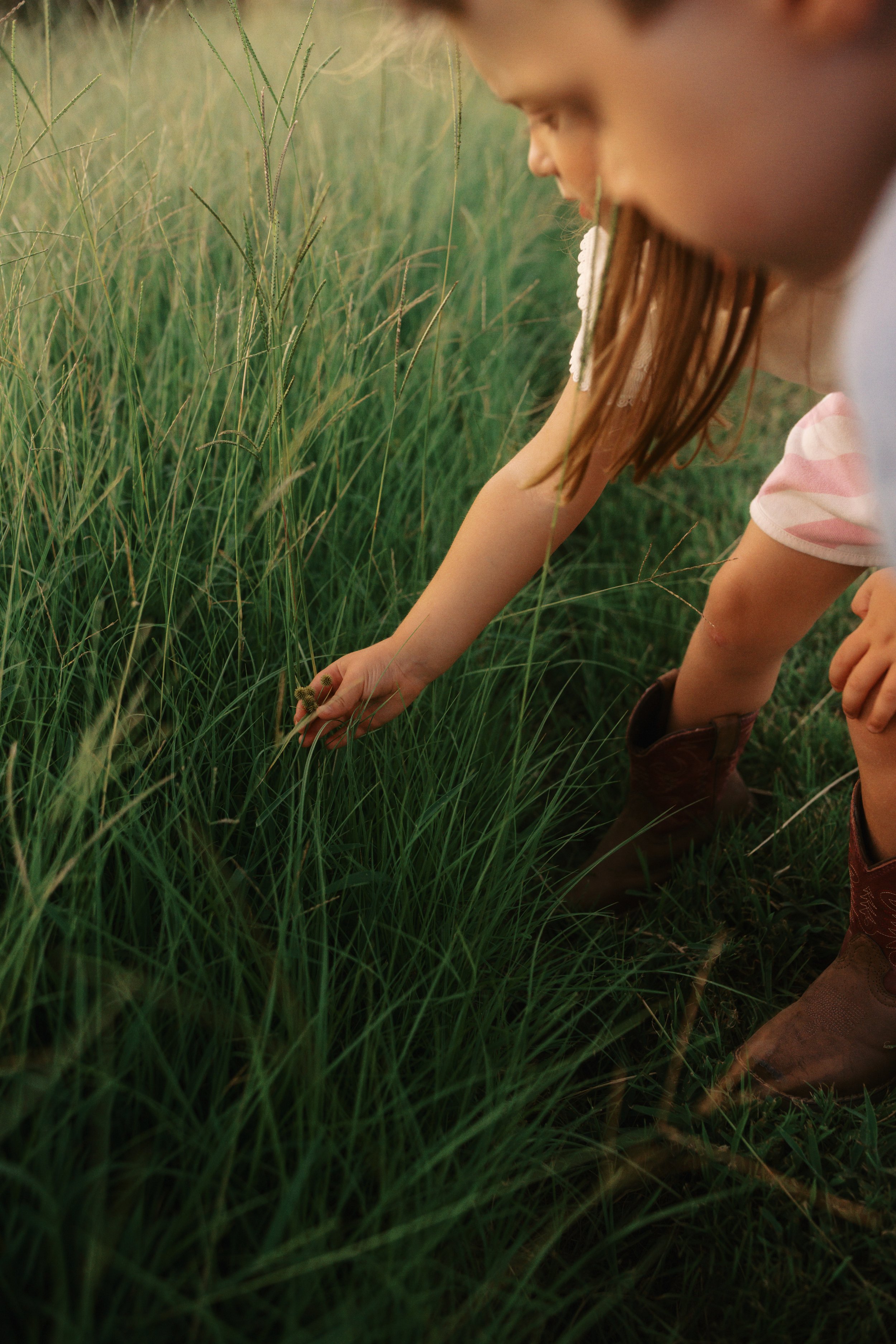 A child crouching in tall green grass, reaching down with one hand, wearing a striped shirt and cowboy boots.