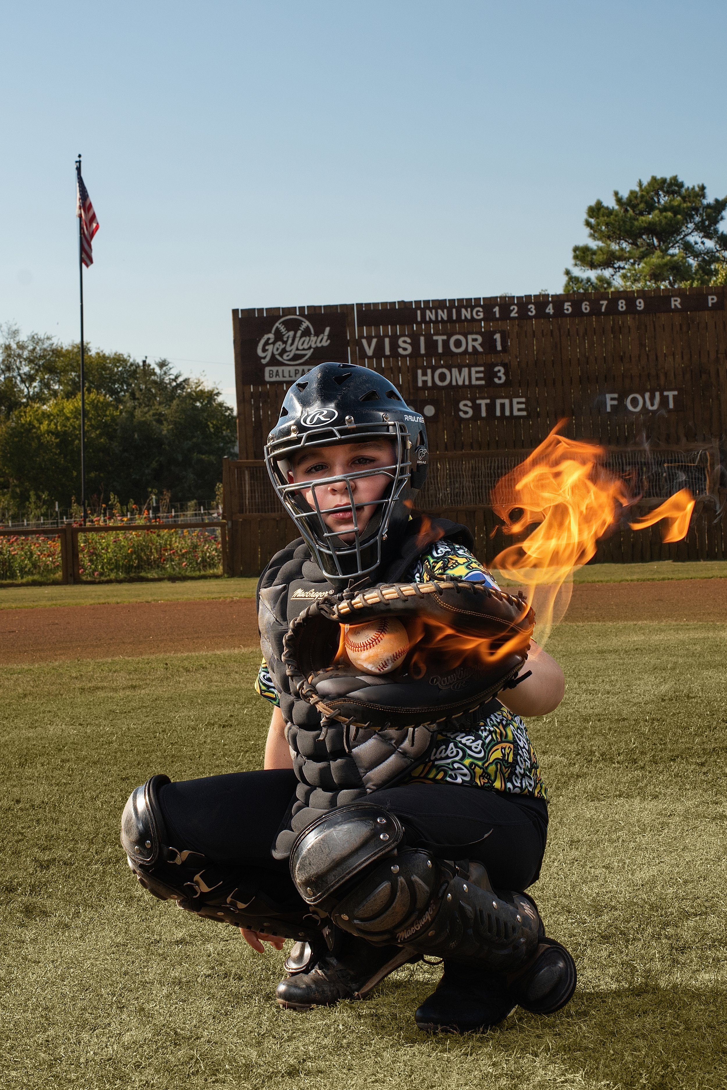 A young boy in baseball catcher's gear, squatting on a baseball field, holding a catcher's mitt with a glowing fireball inside, wearing a helmet and shoulder pads, with a scoreboard and American flag in the background.