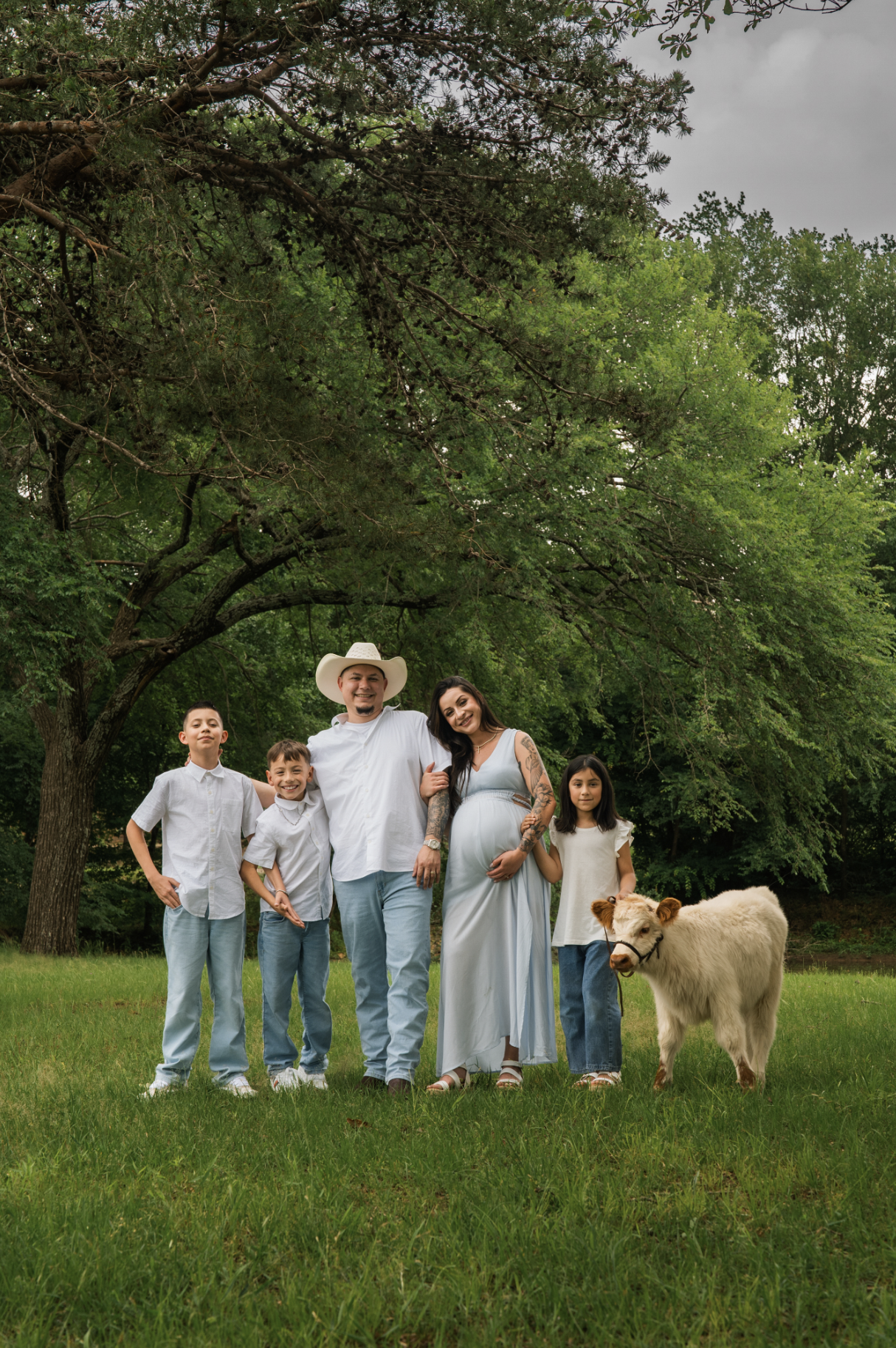A family of five, including three children, an adult man, an adult woman who is pregnant, and a small cow, standing outdoors on a green lawn under a large tree with green foliage. The sky is overcast.