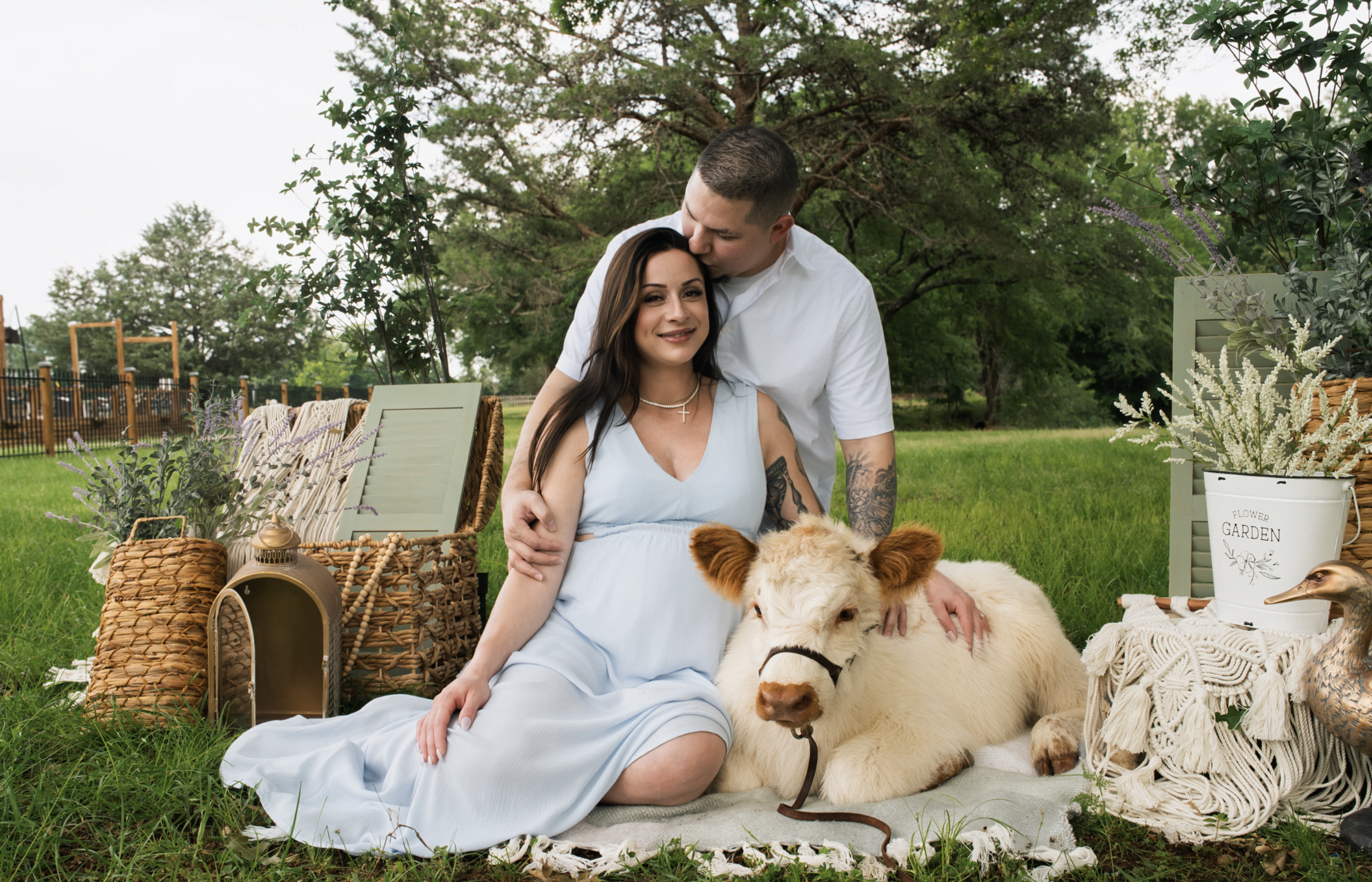 A couple with dogs sitting on a blanket outdoors in a garden, surrounded by plants, baskets, and garden decor.
