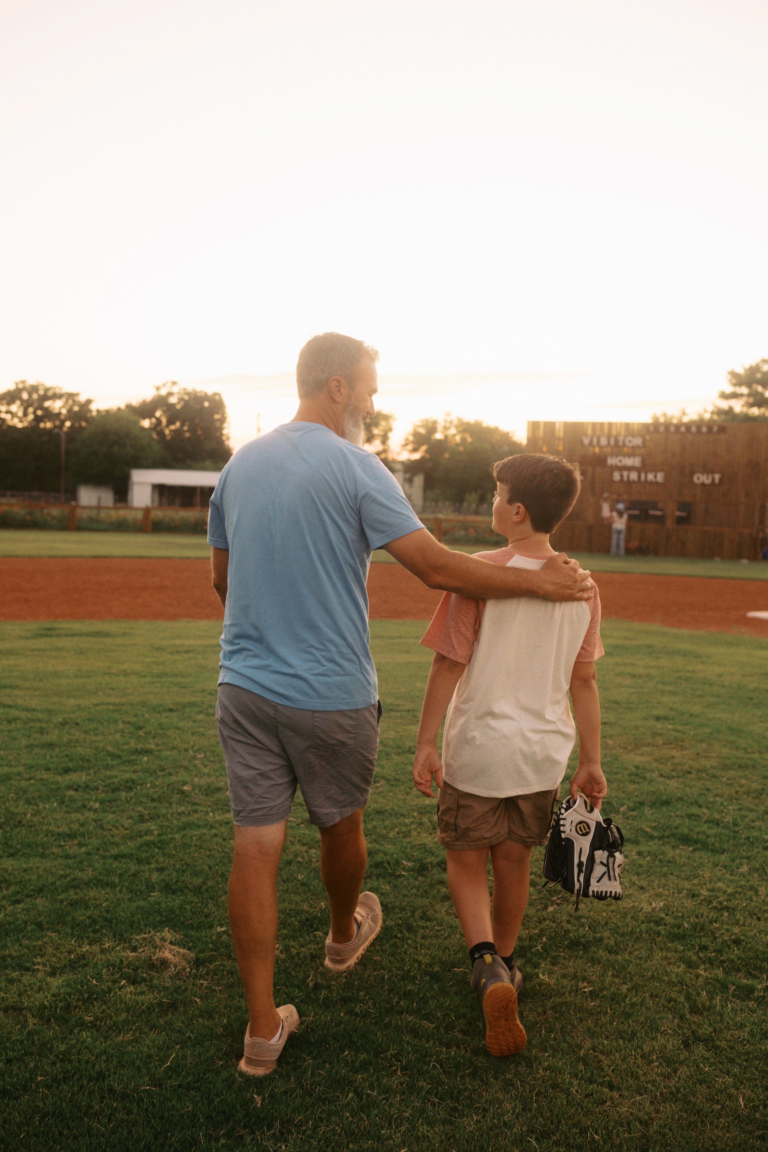 A man and a young boy walking together on a grassy field at sunset, with a baseball scoreboard in the background.