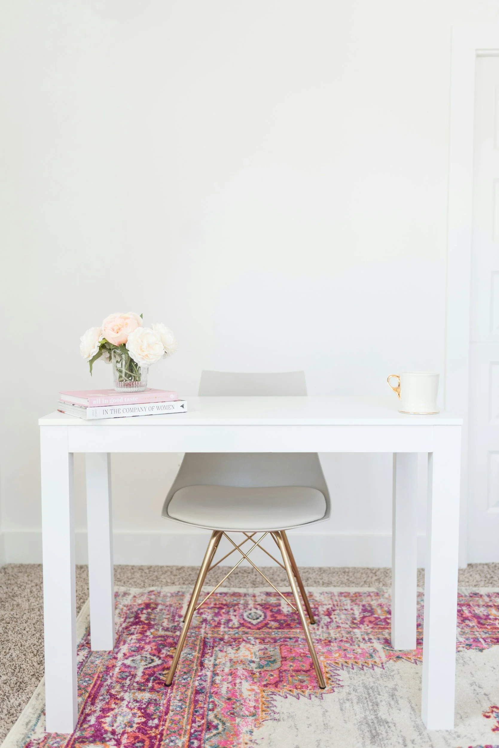 A white desk with a floral arrangement, books, and a cup, set on a multicolored Persian-style rug against a plain white wall.
