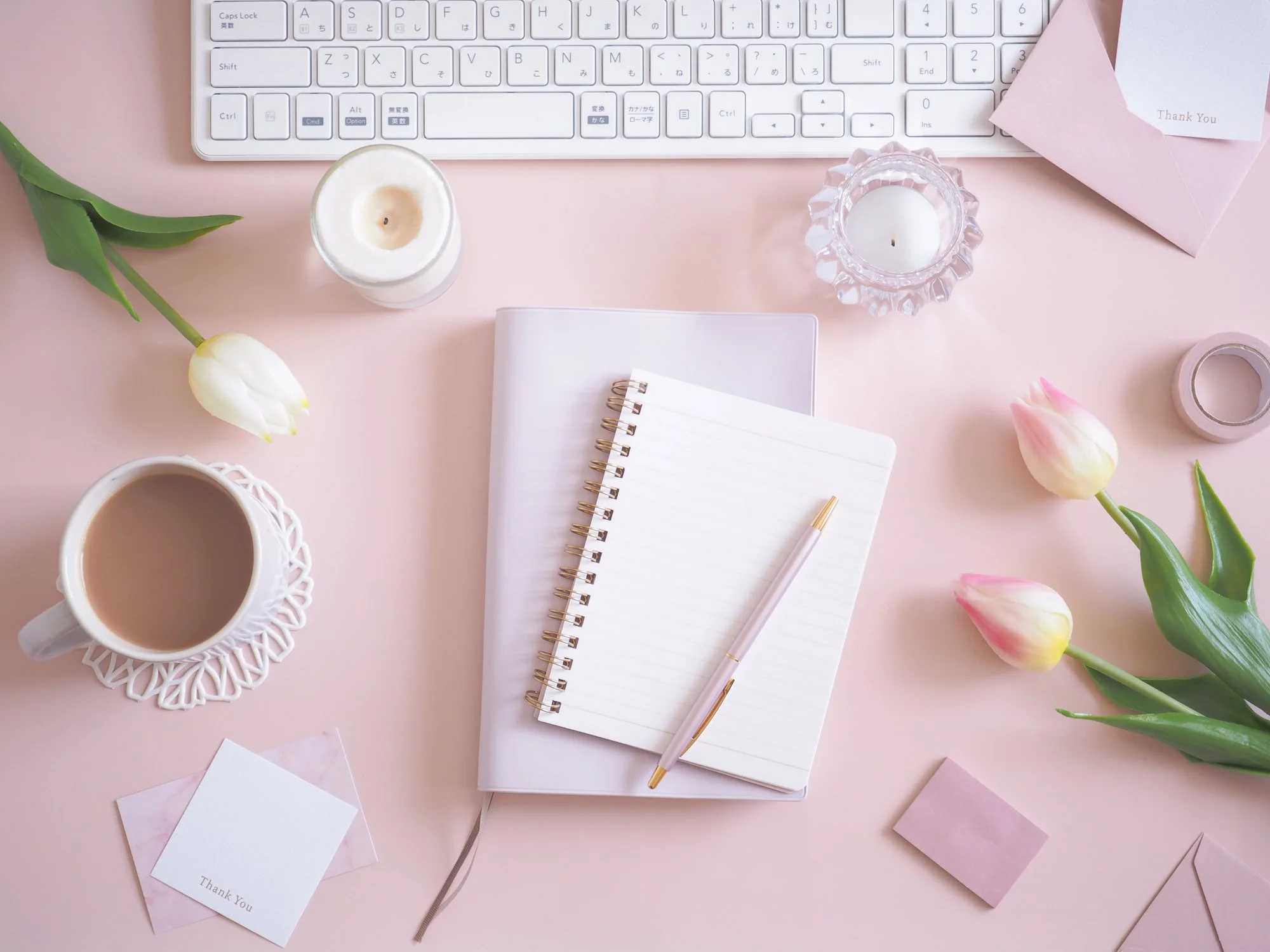 A pink desk with a white keyboard, a notebook, a pen, two tulips, a cup of coffee on a lace coaster, candles, thank you notes, and some pink paper.