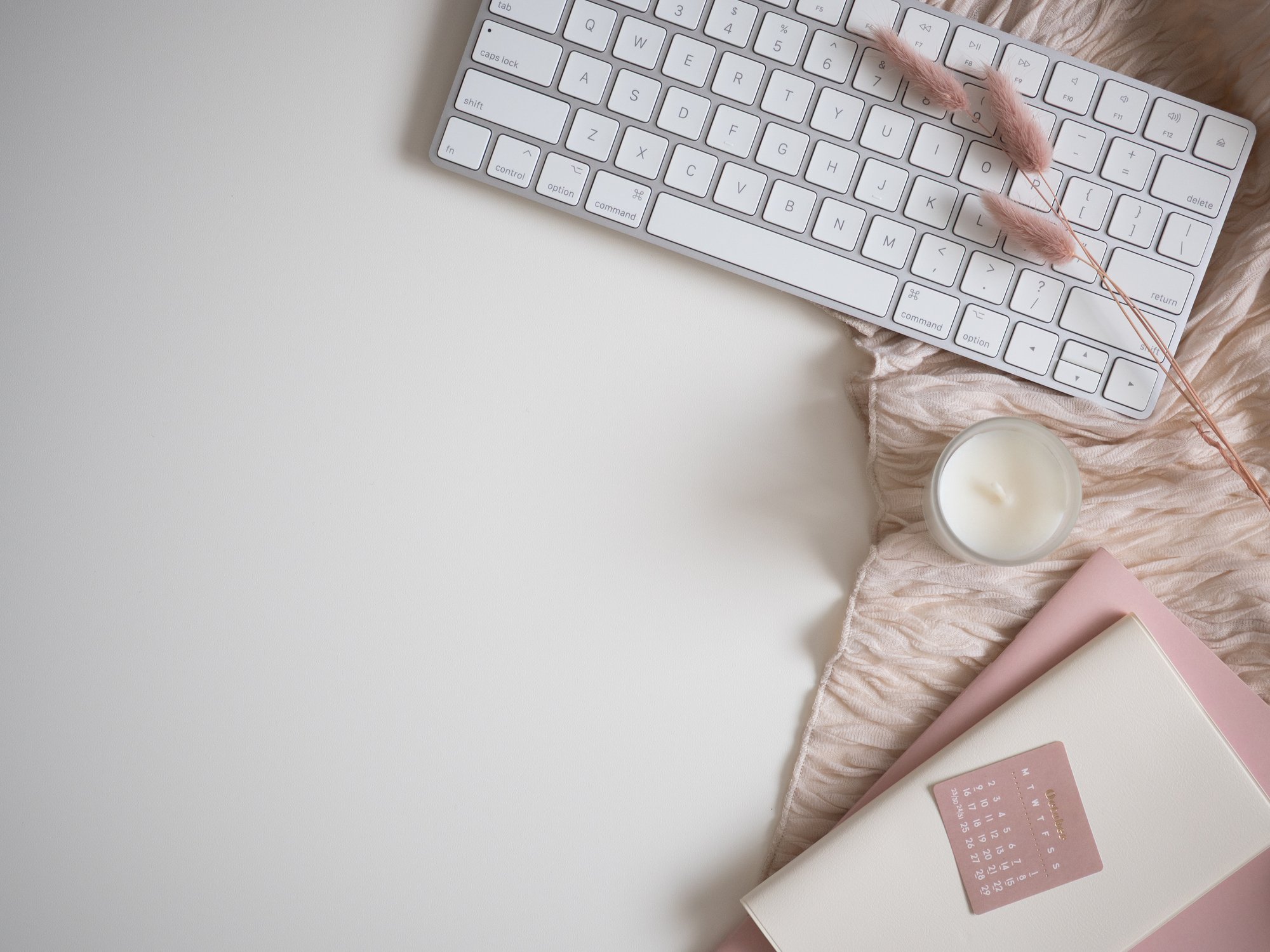 A white computer keyboard with pink fuzzy decorations lying across the top right keys, a lit candle in a glass jar, and a pink notebook on a soft pink cloth on a white surface.