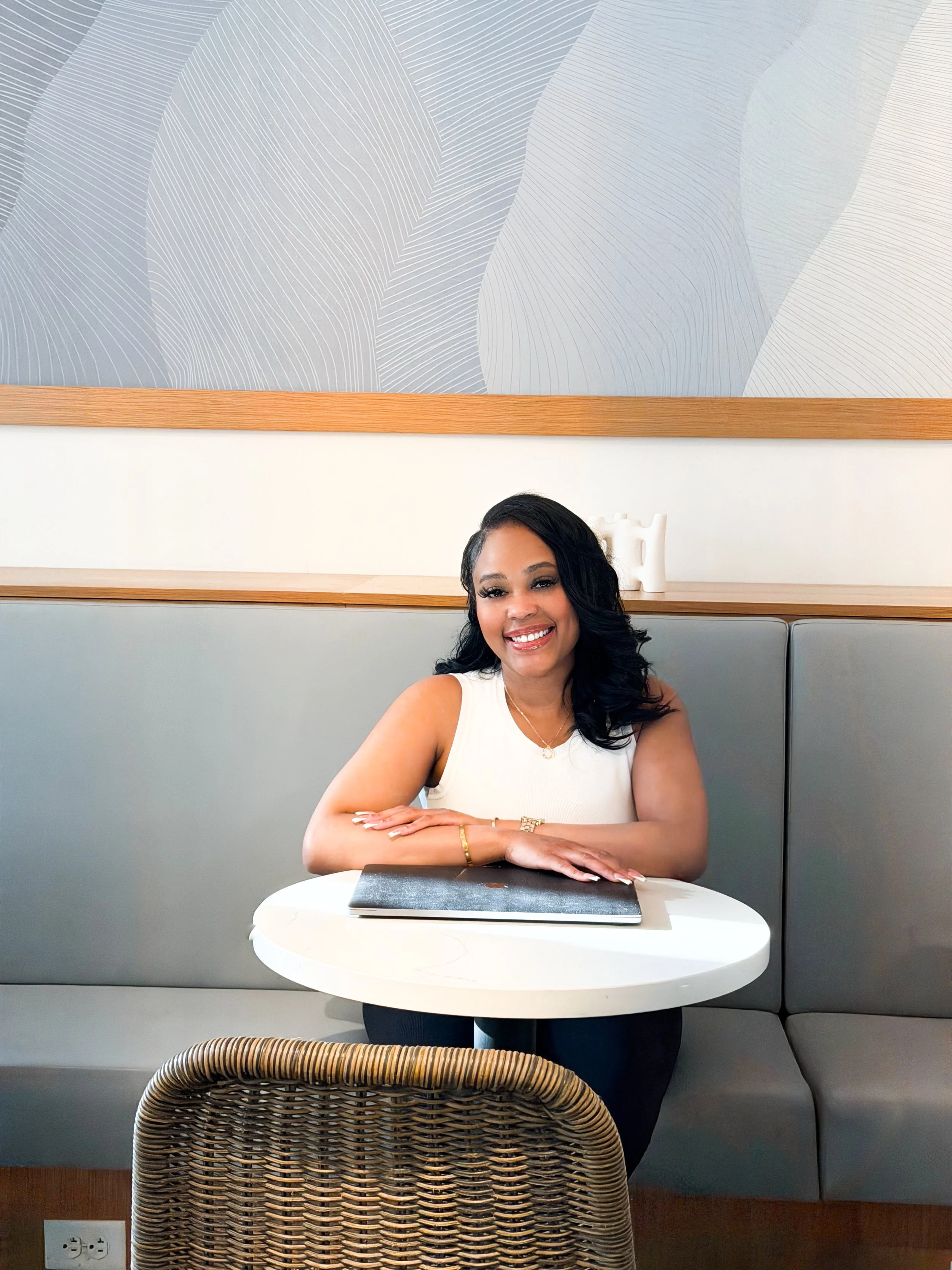 A smiling woman with black hair styled in loose curls, wearing a sleeveless white top, is seated at a small white round table in a modern restaurant or café. She has jewelry on her left wrist and a necklace, and appears to be posing for the photo. There is a gray cushioned seat behind her, a wooden ledge above, and a decorative abstract wall art piece in shades of gray and white hanging on the wall.