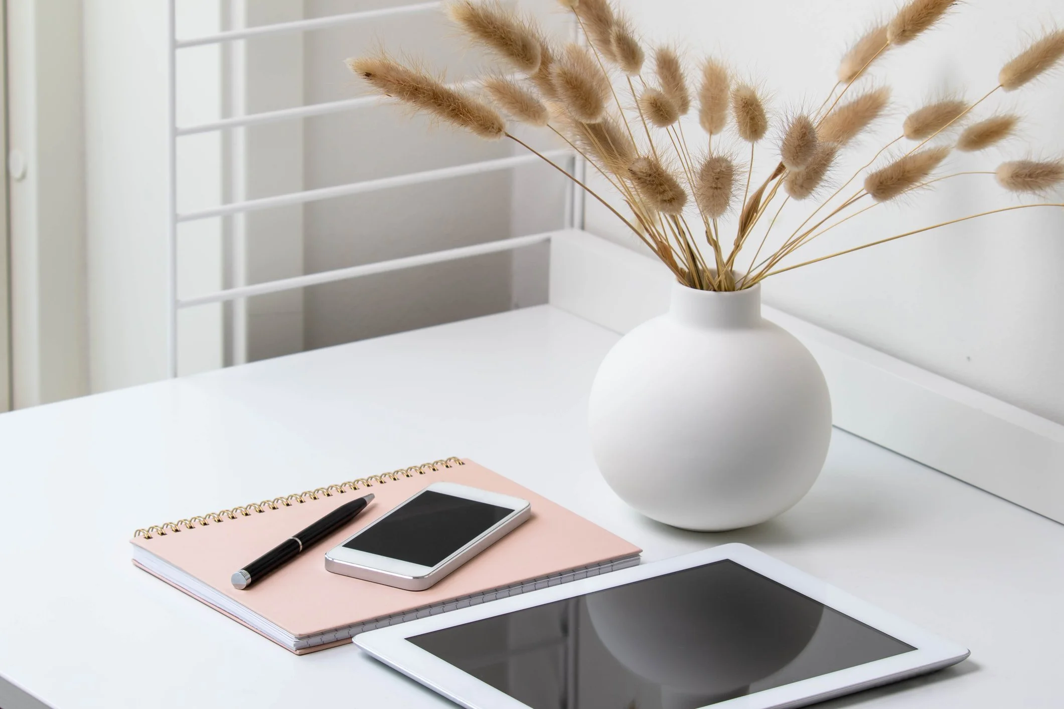White desk with a pink spiral notebook, a black pen, a smartphone, and a tablet. A white vase with beige dried flowers. The background shows a white wall and window blinds.