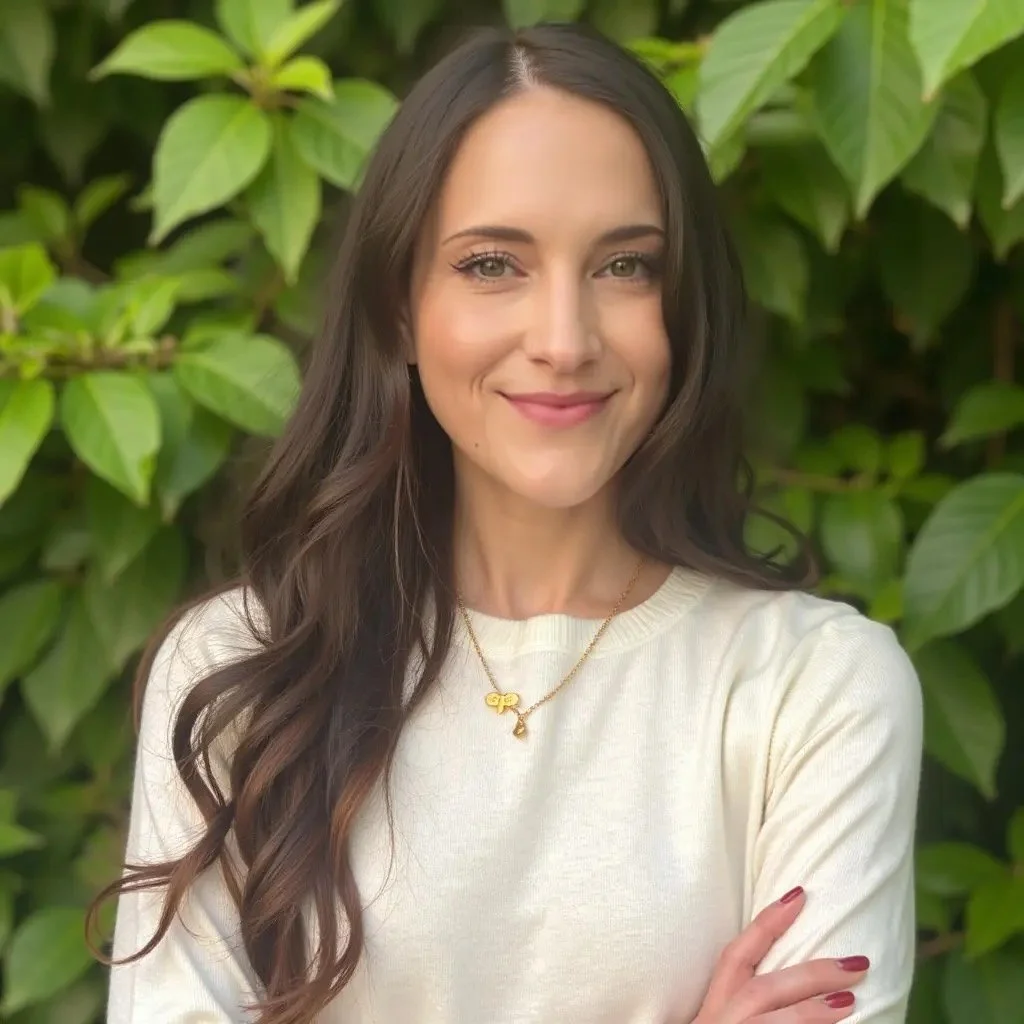A woman with long dark hair smiling outdoors in front of green leaves, wearing a white sweater and a gold necklace.