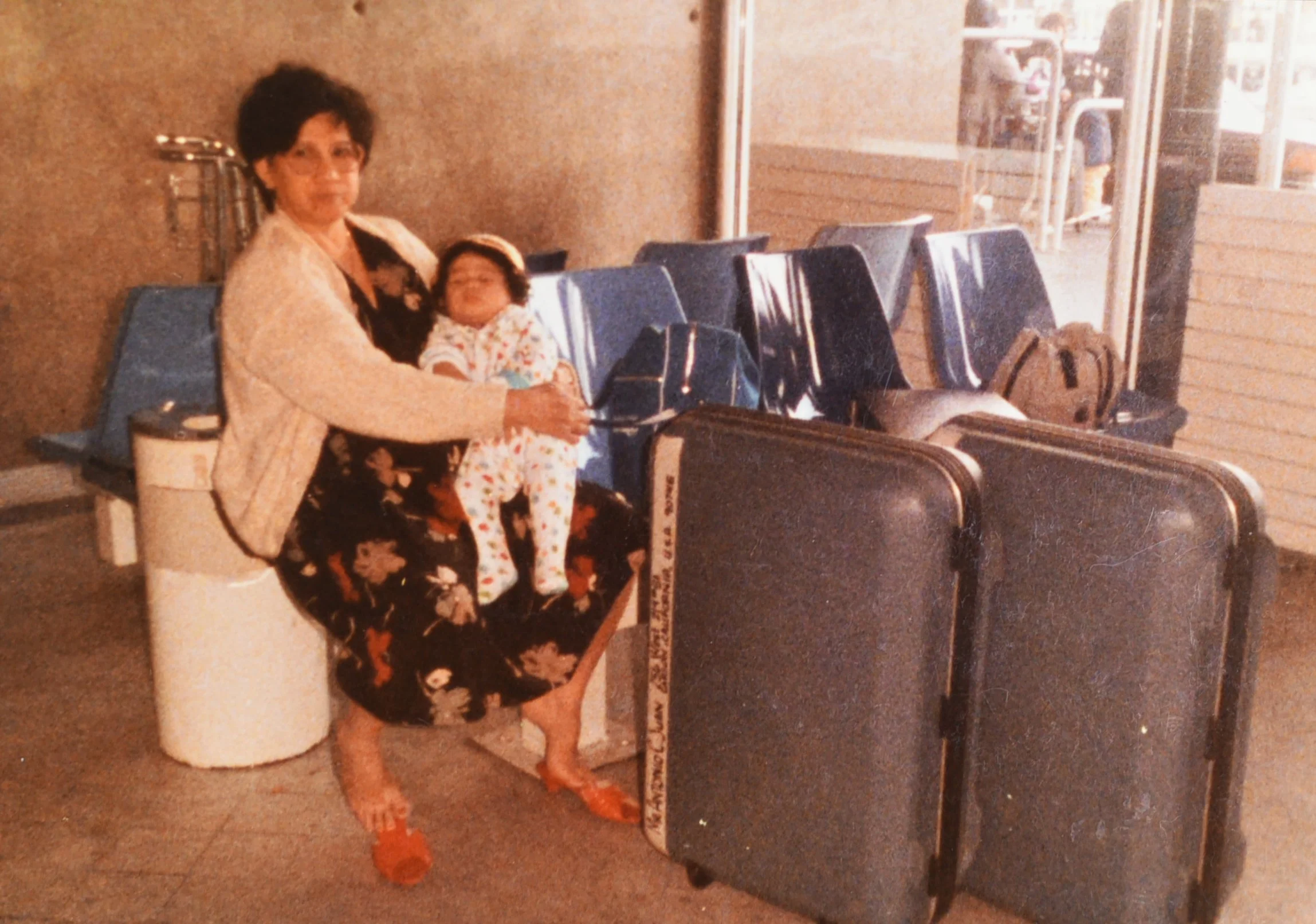 Samantha Juan as a young child sitting on a woman’s lap at an airport in Bahrain, with luggage beside them.