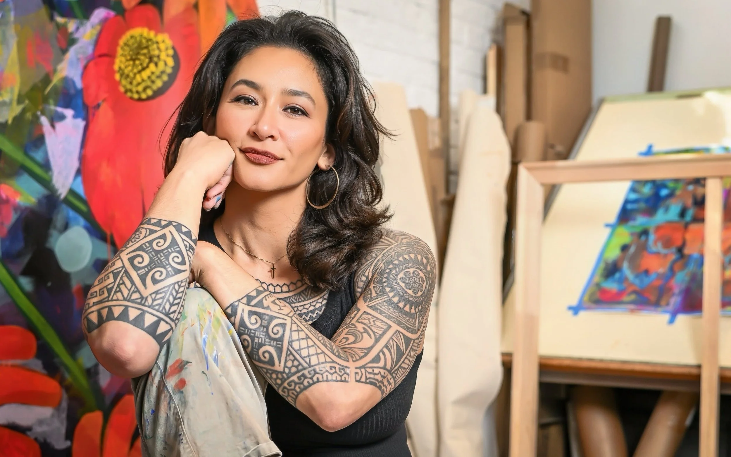 Samantha Juan resting her face on her hand in her art studio, surrounded by colorful paintings and blank canvases.