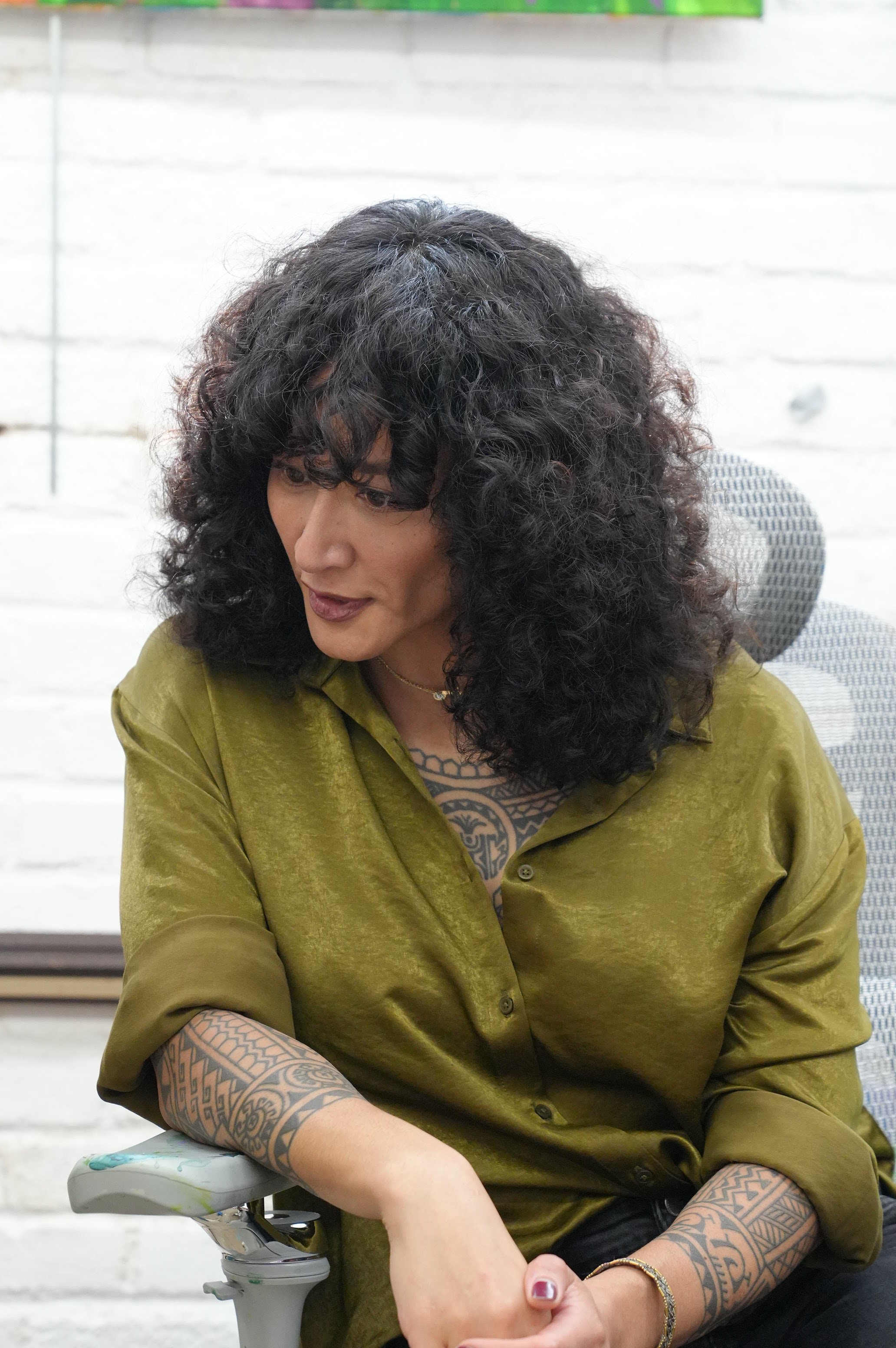 Samantha Juan seated in her studio wearing an olive-green top against a white brick wall.