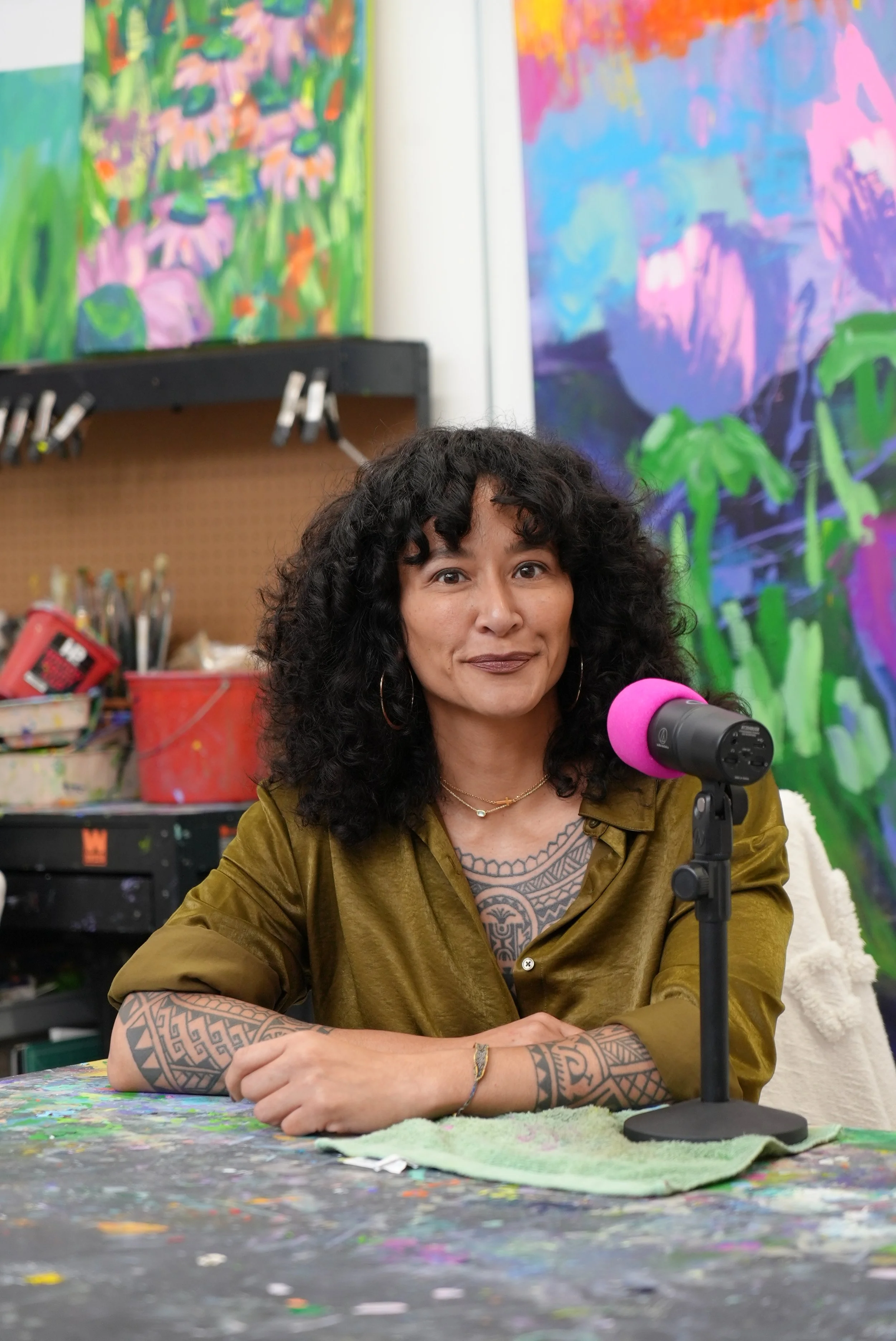 Samantha Juan speaking into a pink microphone while seated in her art studio, surrounded by paintings and art supplies.