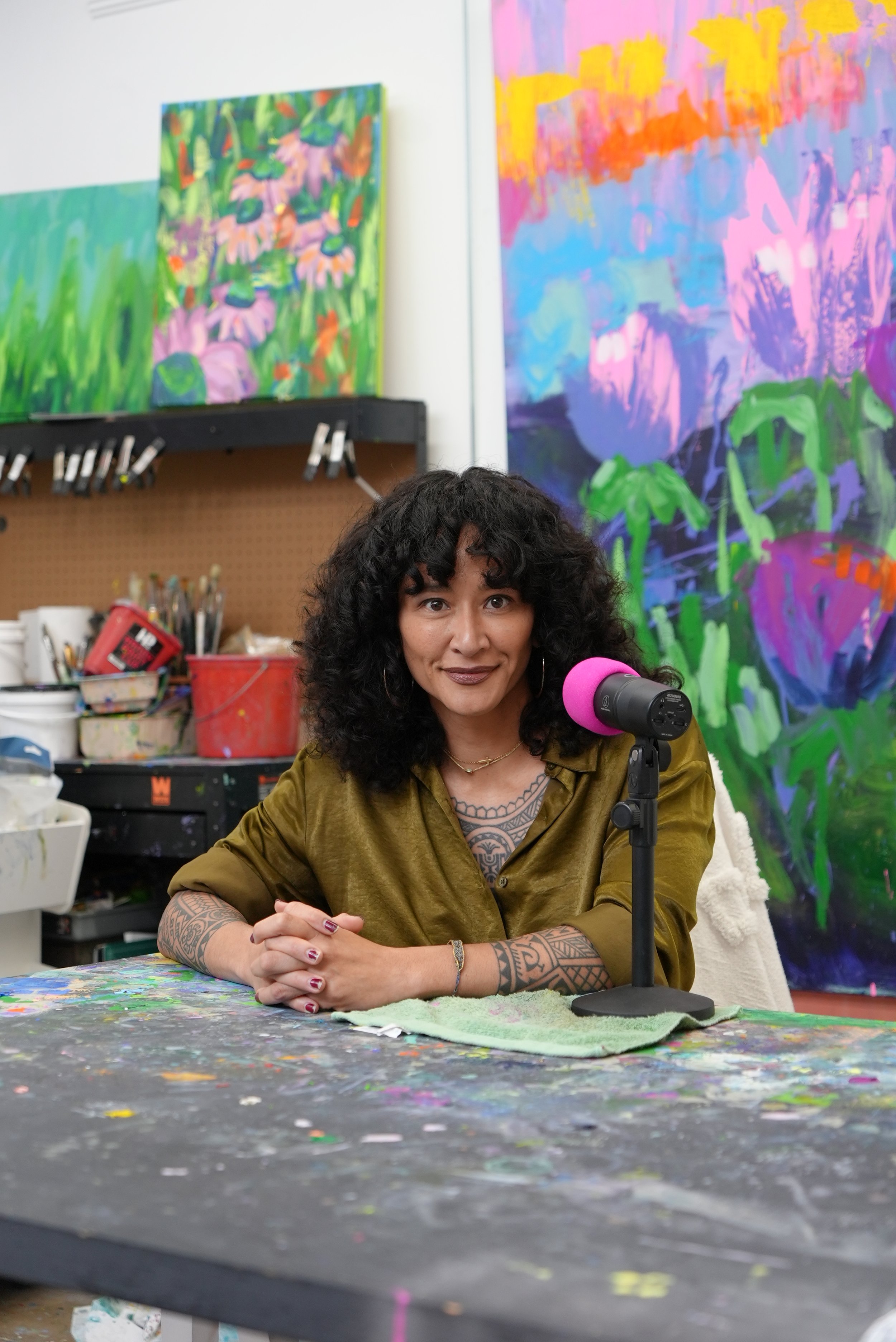 Samantha Juan recording a podcast interview at a paint-covered table surrounded by her colorful abstract artwork.