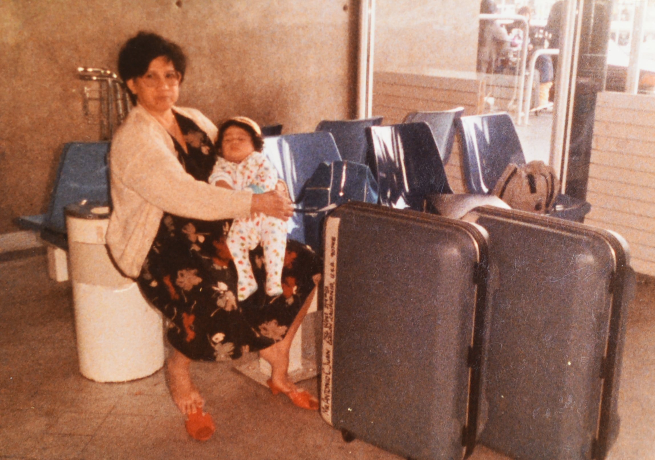 Samantha Juan as a young child sitting on a woman’s lap at an airport in Bahrain, with luggage beside them.