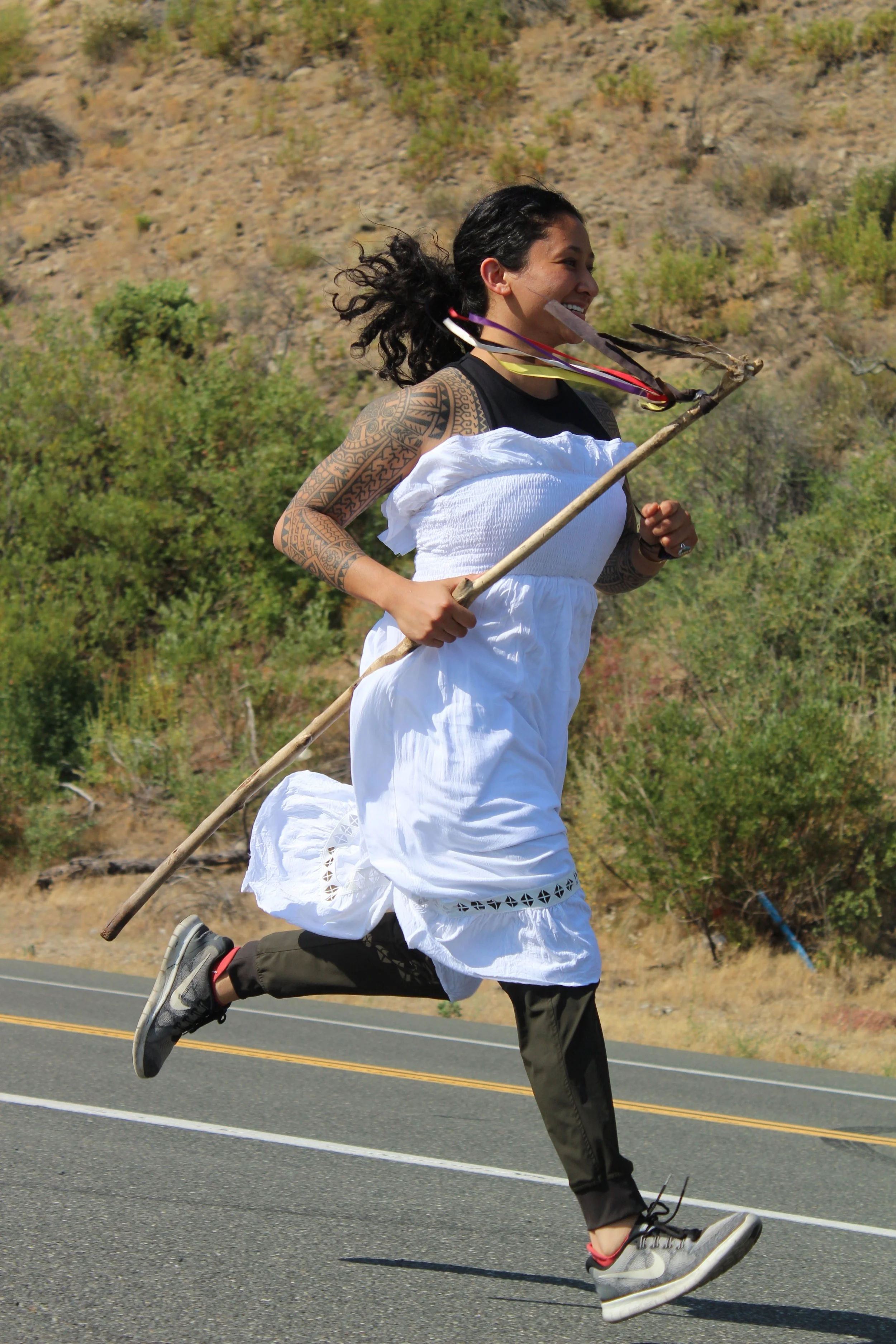 A woman with long, curly hair and a tattooed arm is running outdoors on a road with a scenic hillside and greenery in the background. She is smiling, dressed in a white dress, and holding a stick with feathers and ribbons attached. She is wearing gray sneakers.
