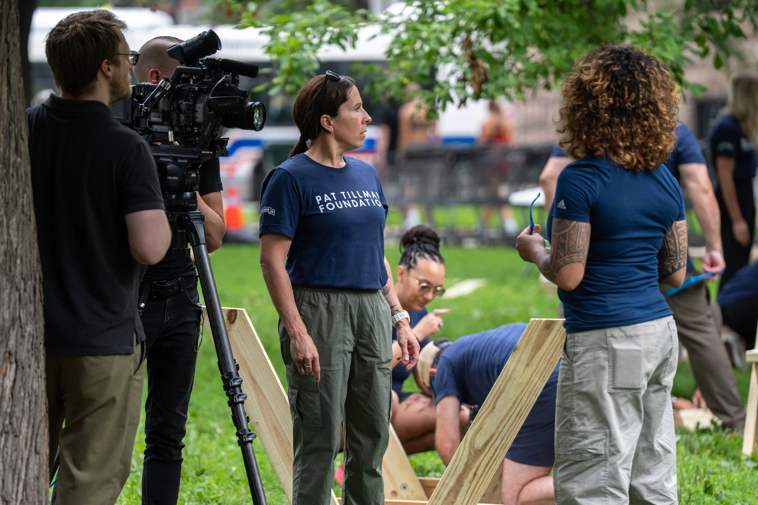 Samantha Juan speaking with another participant during an outdoor community event, with volunteers and wooden materials in the background.