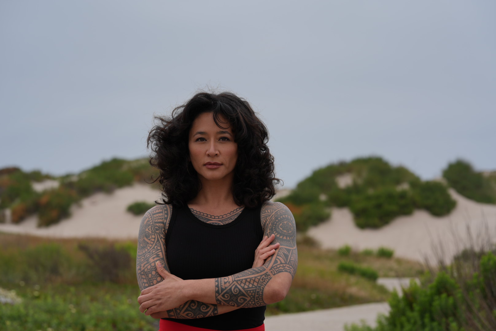 Samantha Juan standing with her arms crossed near sand dunes and coastal greenery.
