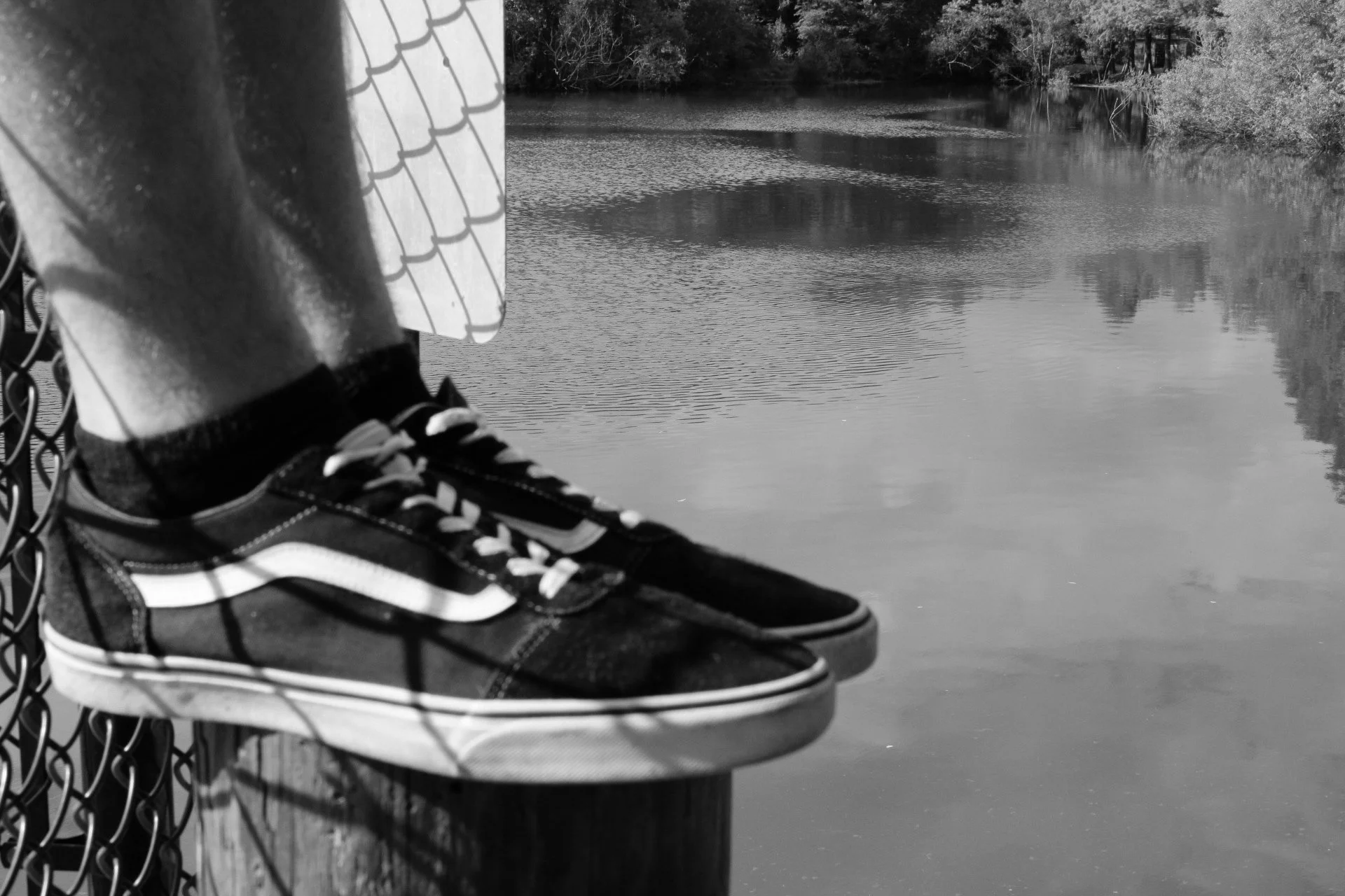 Black and white photo of a person's foot wearing a sneaker resting on a wooden bench near a body of water with trees in the background.