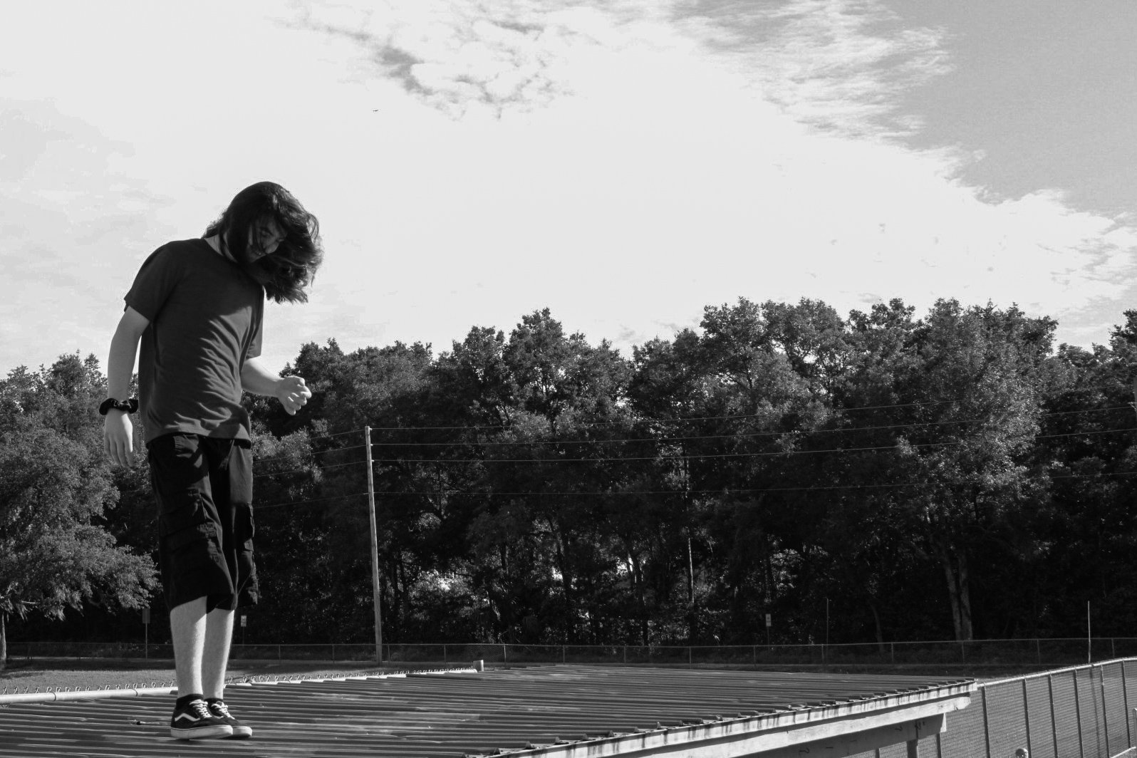 A black and white photo of a young person with shoulder-length hair, wearing a t-shirt, shorts, and sneakers, standing on a sports track or field. They are looking down at the ground with a slightly tilted posture, with trees and a partly cloudy sky in the background.