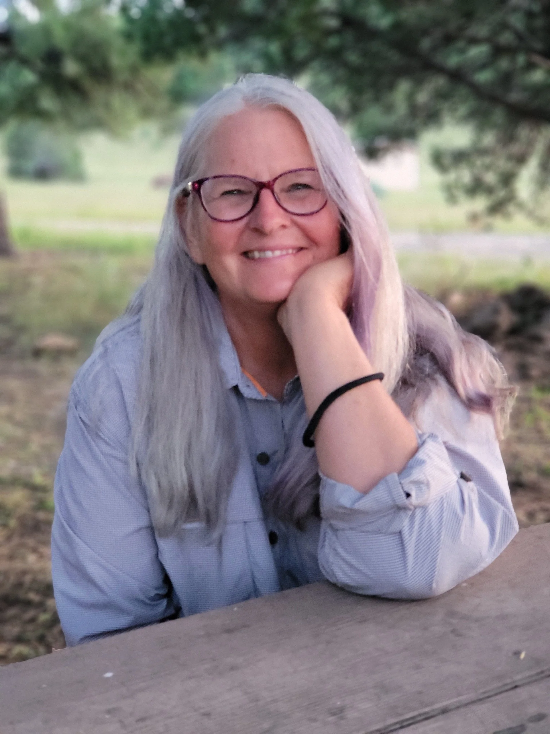 A woman with long gray hair wearing glasses, smiling, resting her chin on her hand, sitting outdoors near a wooden surface, with trees and grass in the background.