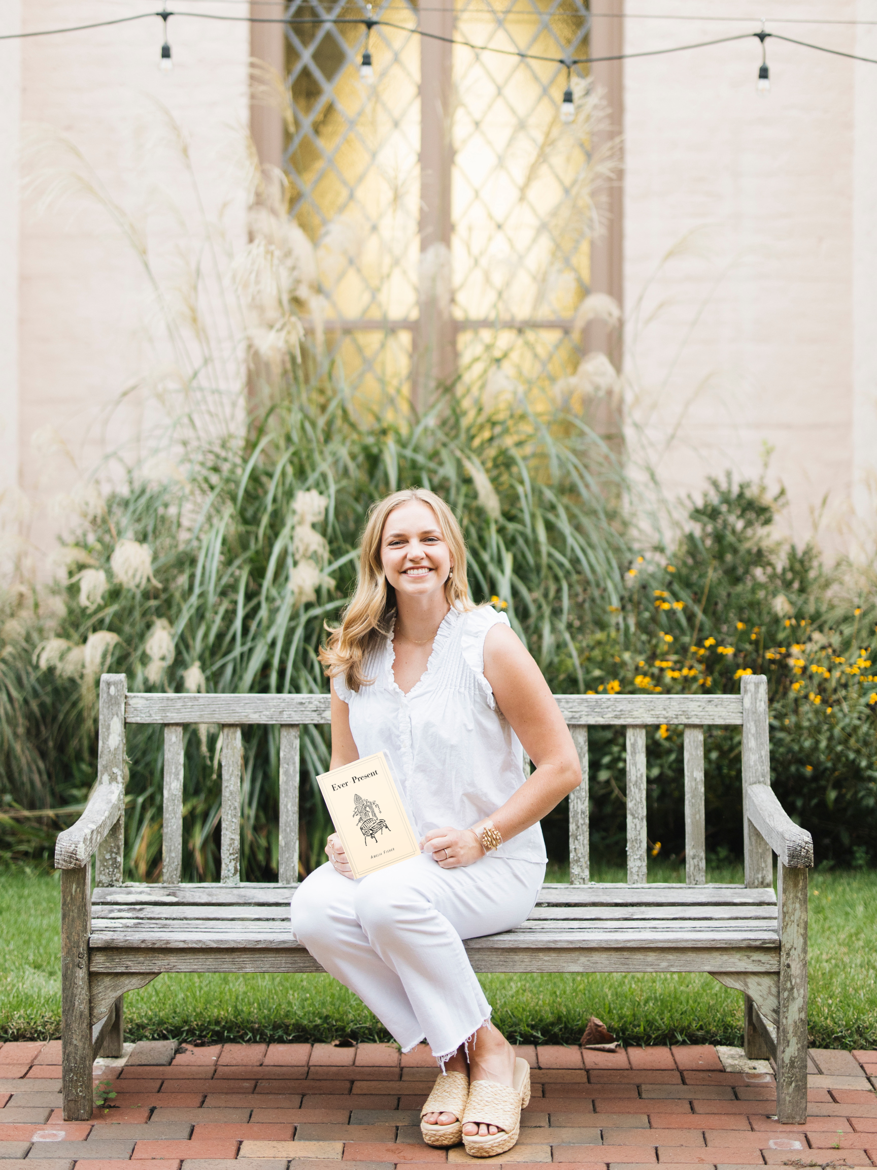 A young woman sitting on a wooden bench outdoors, holding a book, smiling, with plants and a decorative background behind her.