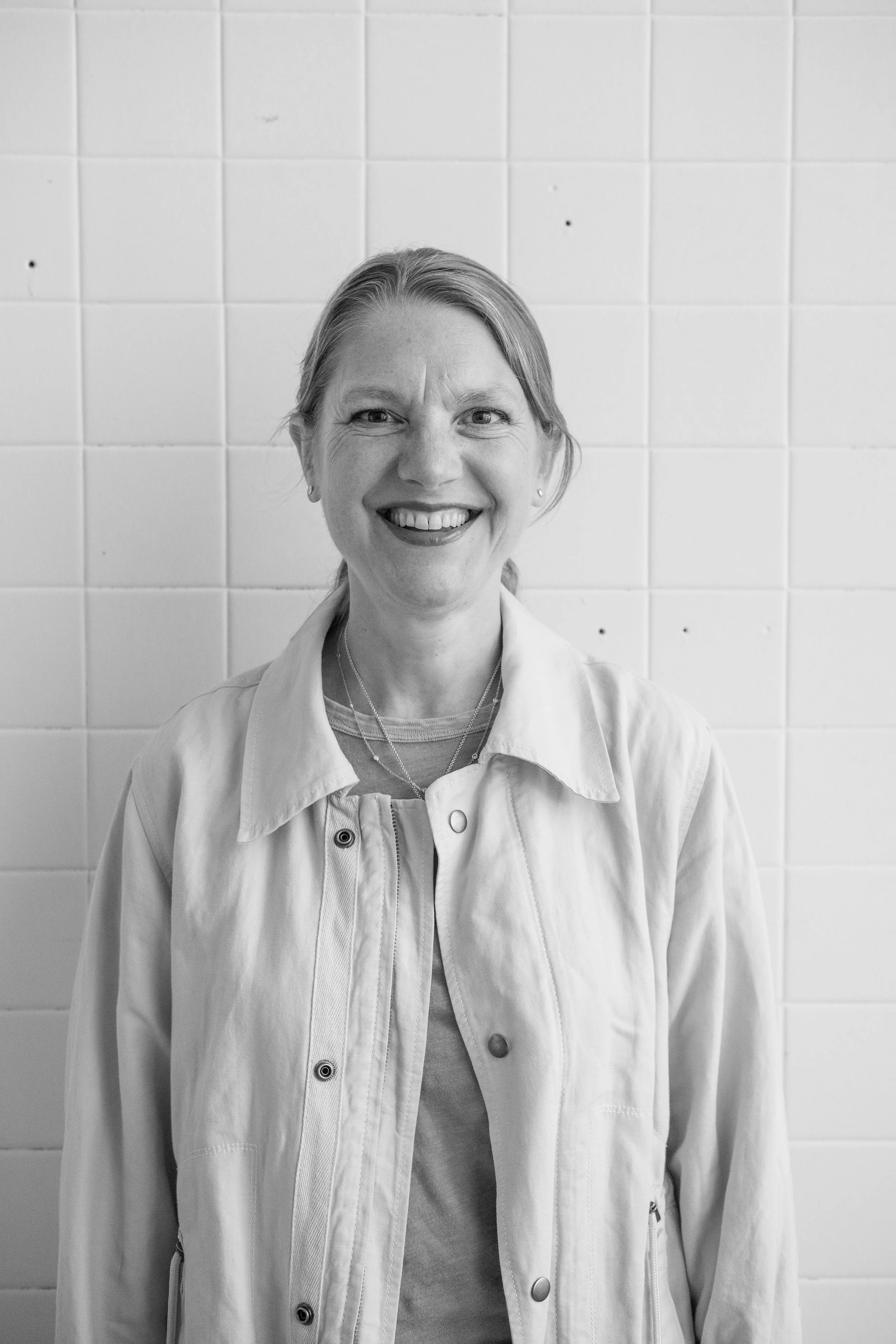 A woman smiling, wearing a light-colored jacket and a shirt, standing in front of a white tiled wall.