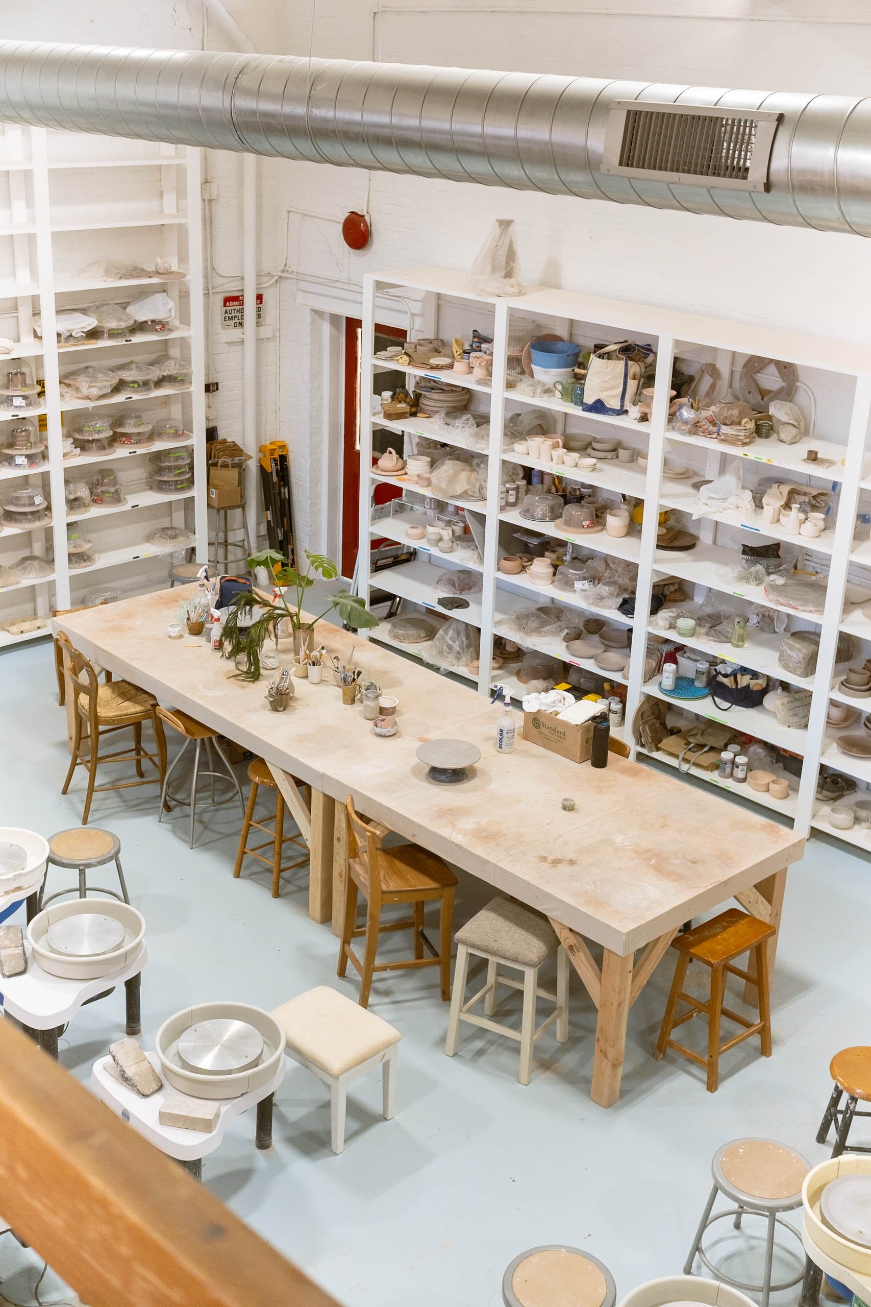 Image of a pottery or ceramic workshop with white shelves filled with various pottery pieces, a large wooden work table with pottery supplies, and numerous stools.