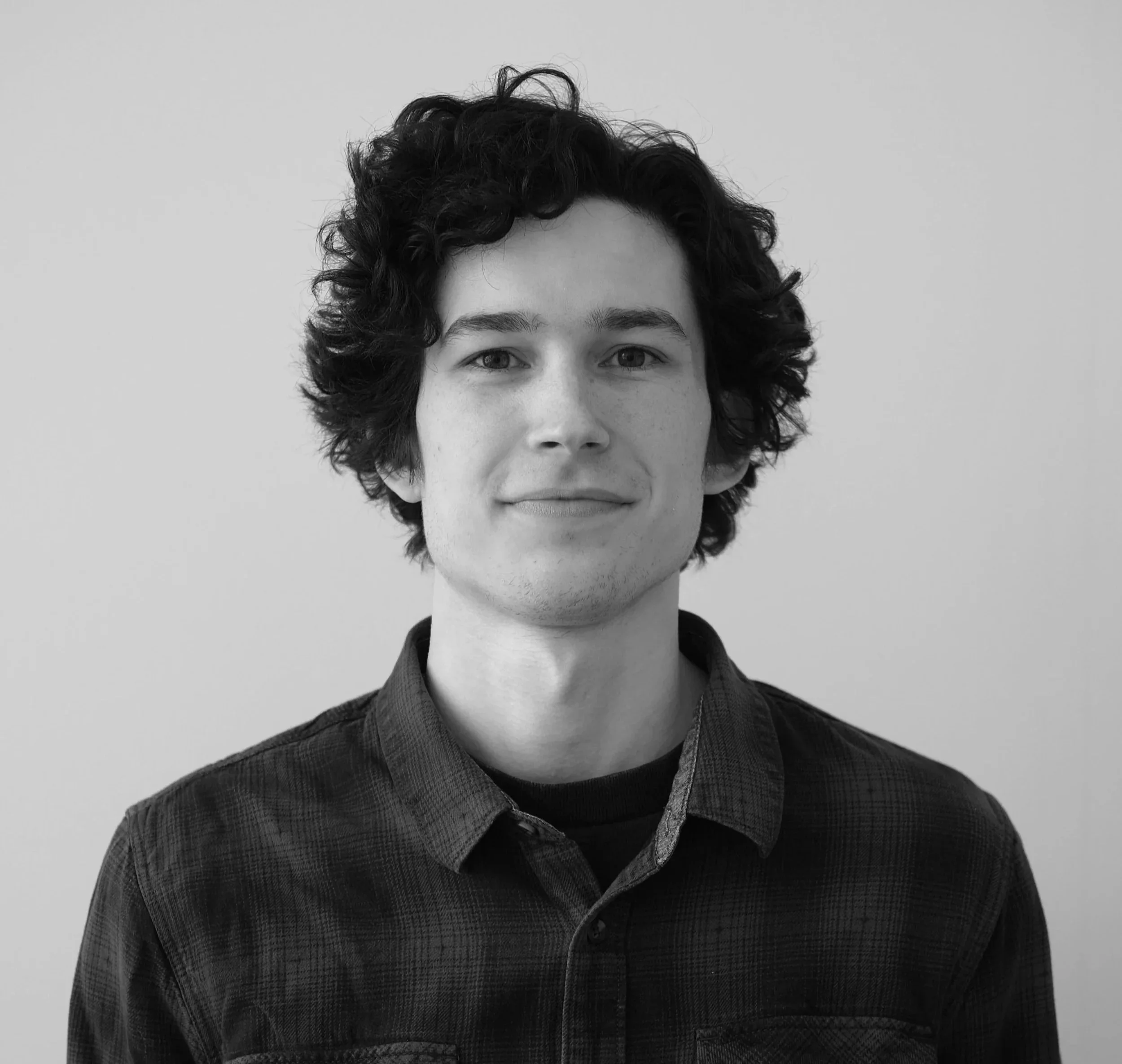 Close-up of a young person with curly hair, wearing a dark collared shirt with a subtle checkered pattern, smiling slightly against a plain background.