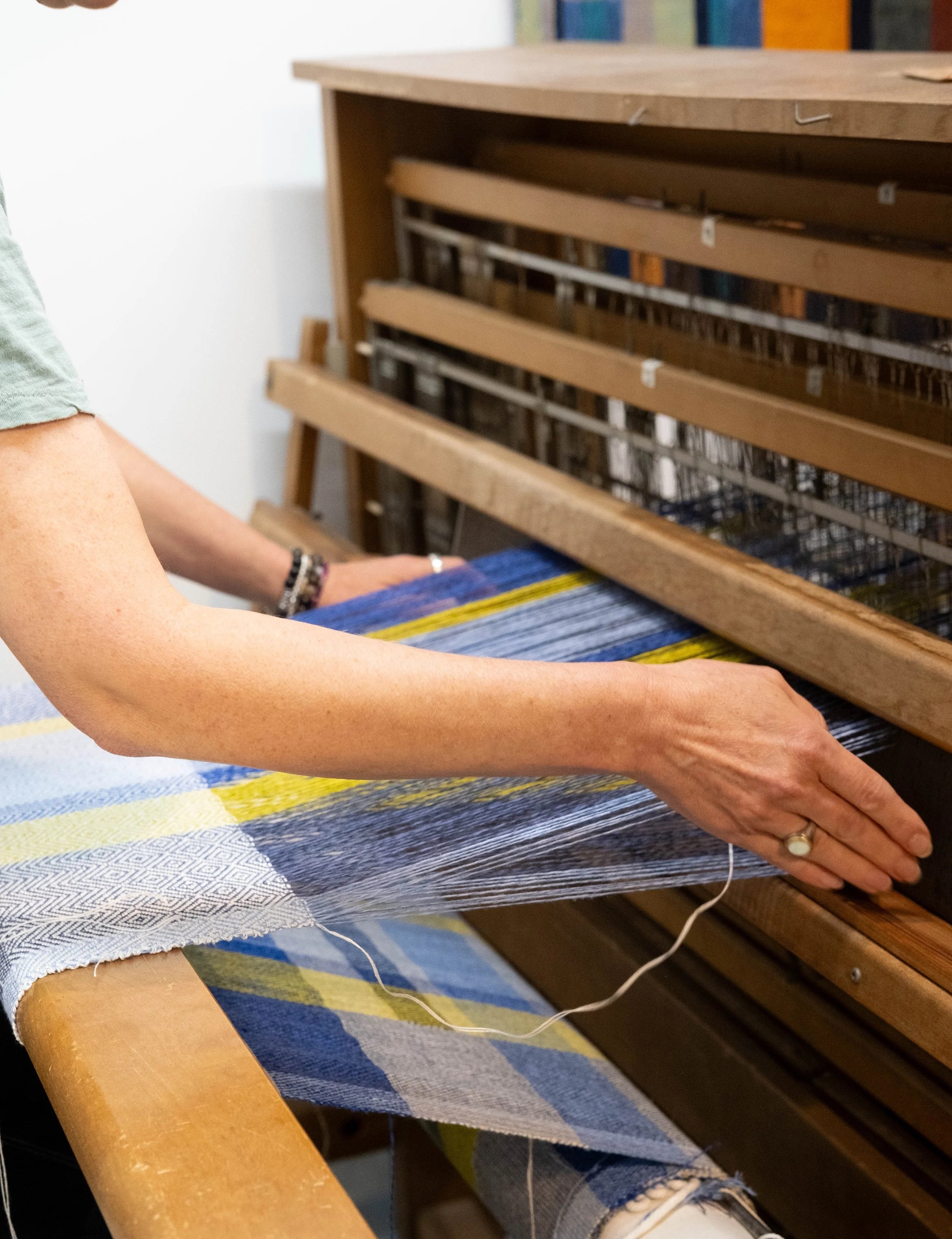 Person weaving fabric on a loom with colorful yarns and a multicolored woven cloth. The person's arm is visible, wearing a ring on the finger and a beaded bracelet.