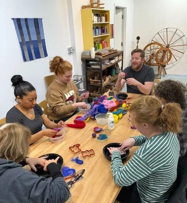 Group of six people sitting around a wooden table, engaged in a craft activity with colorful felt and wool, in a cozy room with shelves and a spinning wheel in the background.