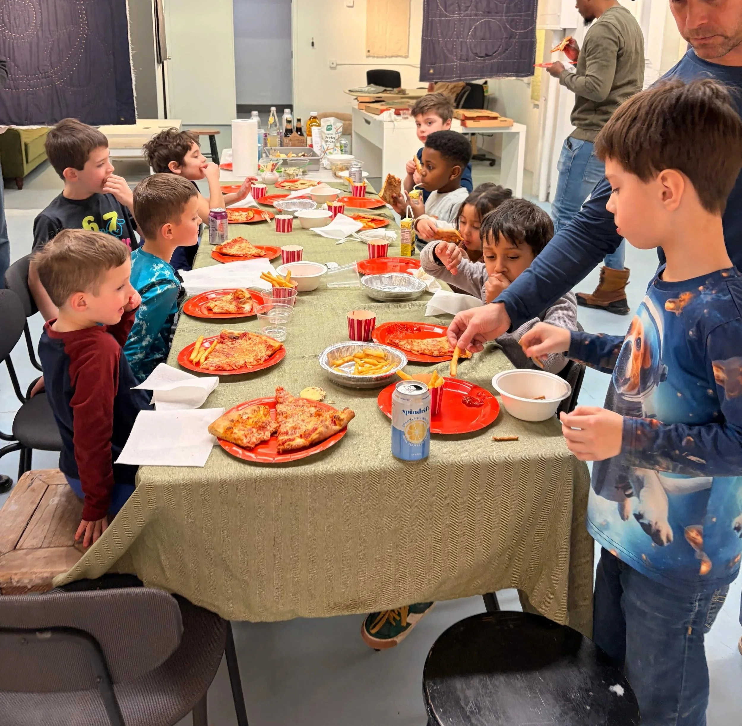 Children sitting at a long table with pizza, fries, and drinks during a gathering. Adults are standing nearby, some eating and others preparing food or cleaning up.