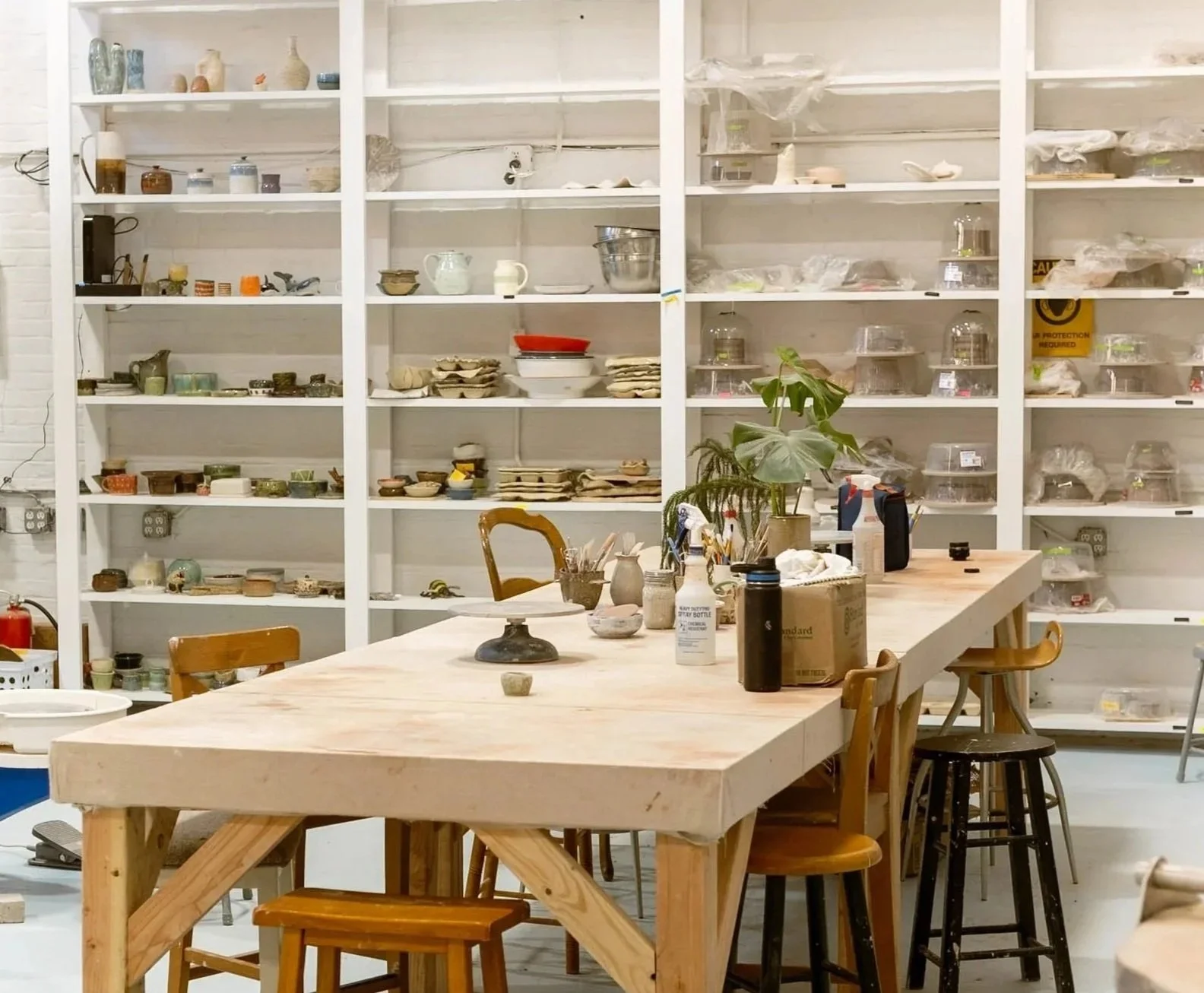 A studio or workshop space featuring a large wooden table with chairs, various art supplies, and shelves filled with ceramics, pottery, and art tools. There is a plant on the table and some items in spray bottles and boxes.