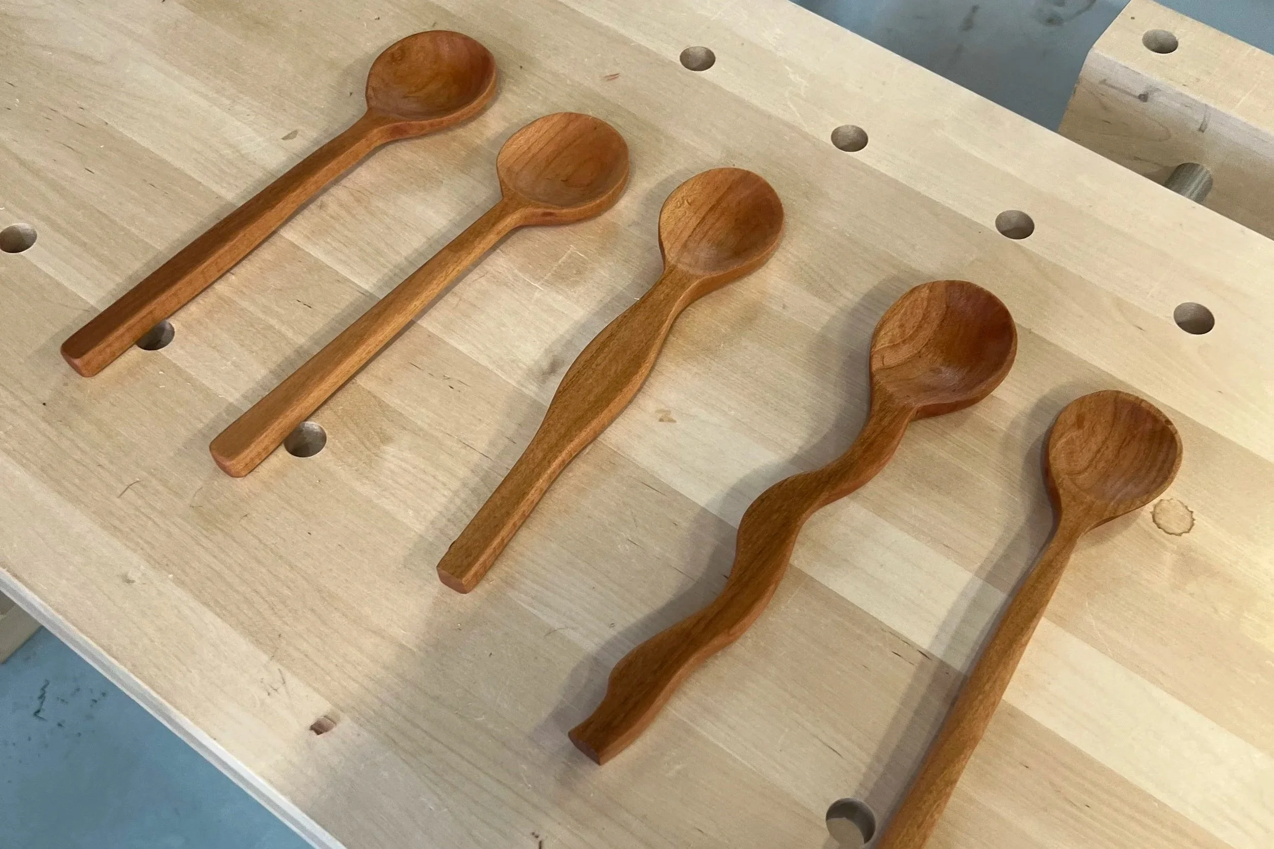 Four wooden spoons placed in a row on a wooden workbench with evenly spaced holes.