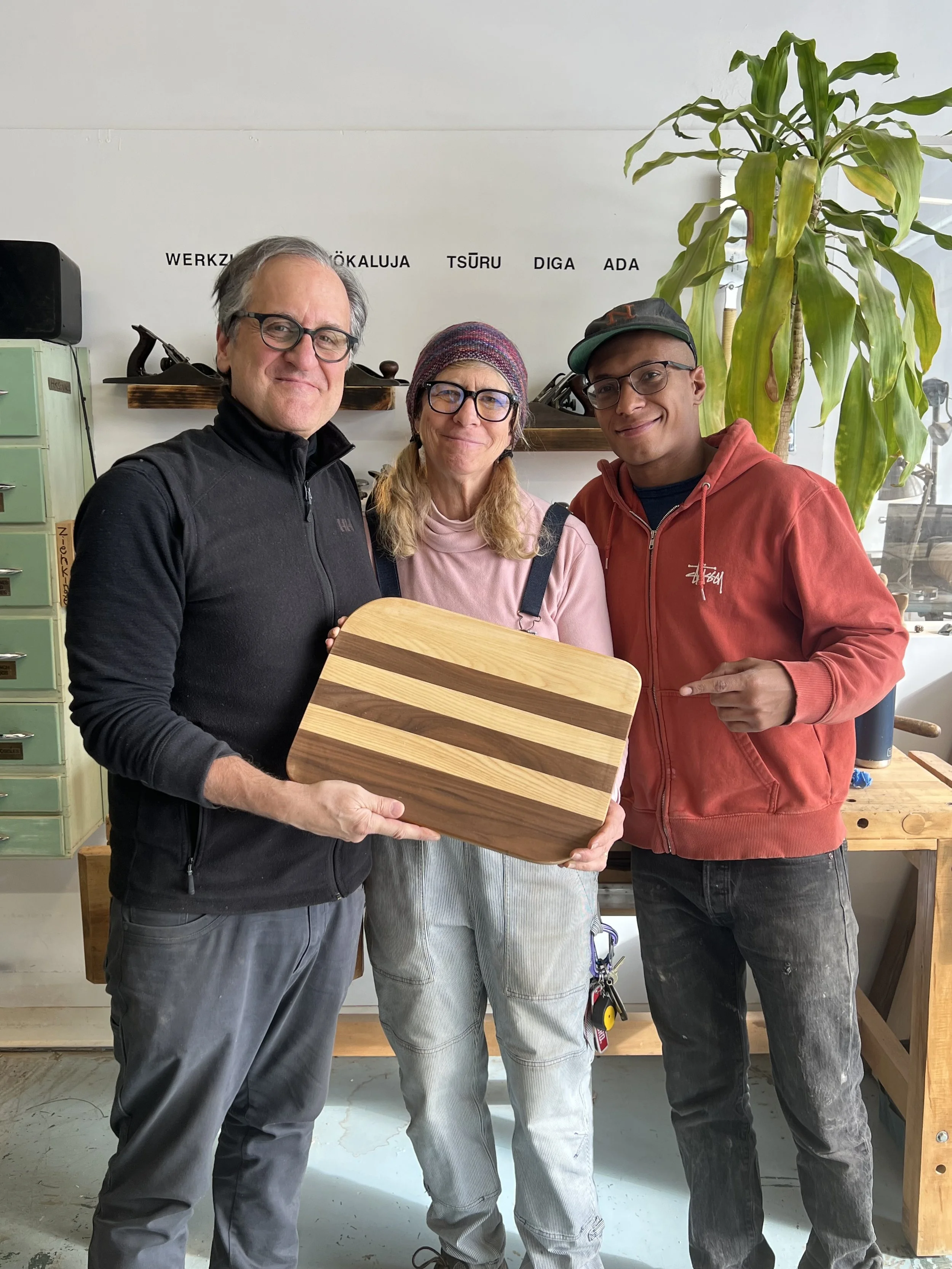 Three people in a workshop holding a striped wooden cutting board, with tools on shelves and a large leafy plant in the background.