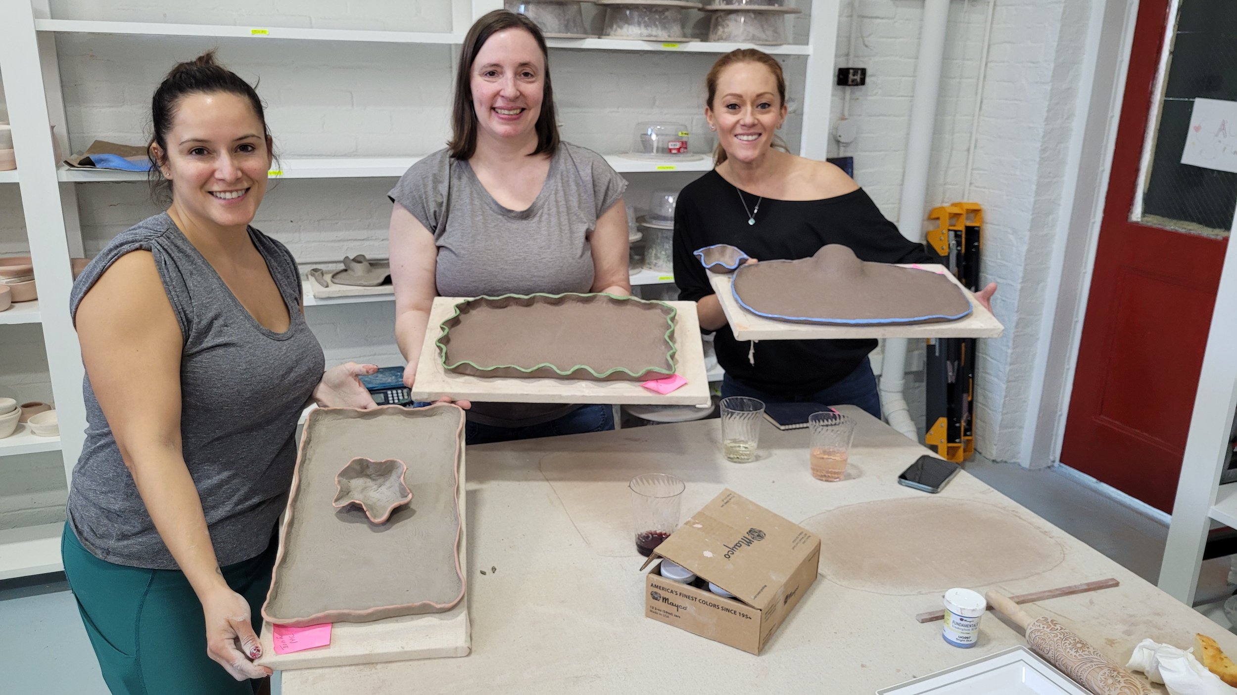 Three women smiling at a pottery workshop, holding painted ceramic trays, with various pottery tools and supplies on the table.