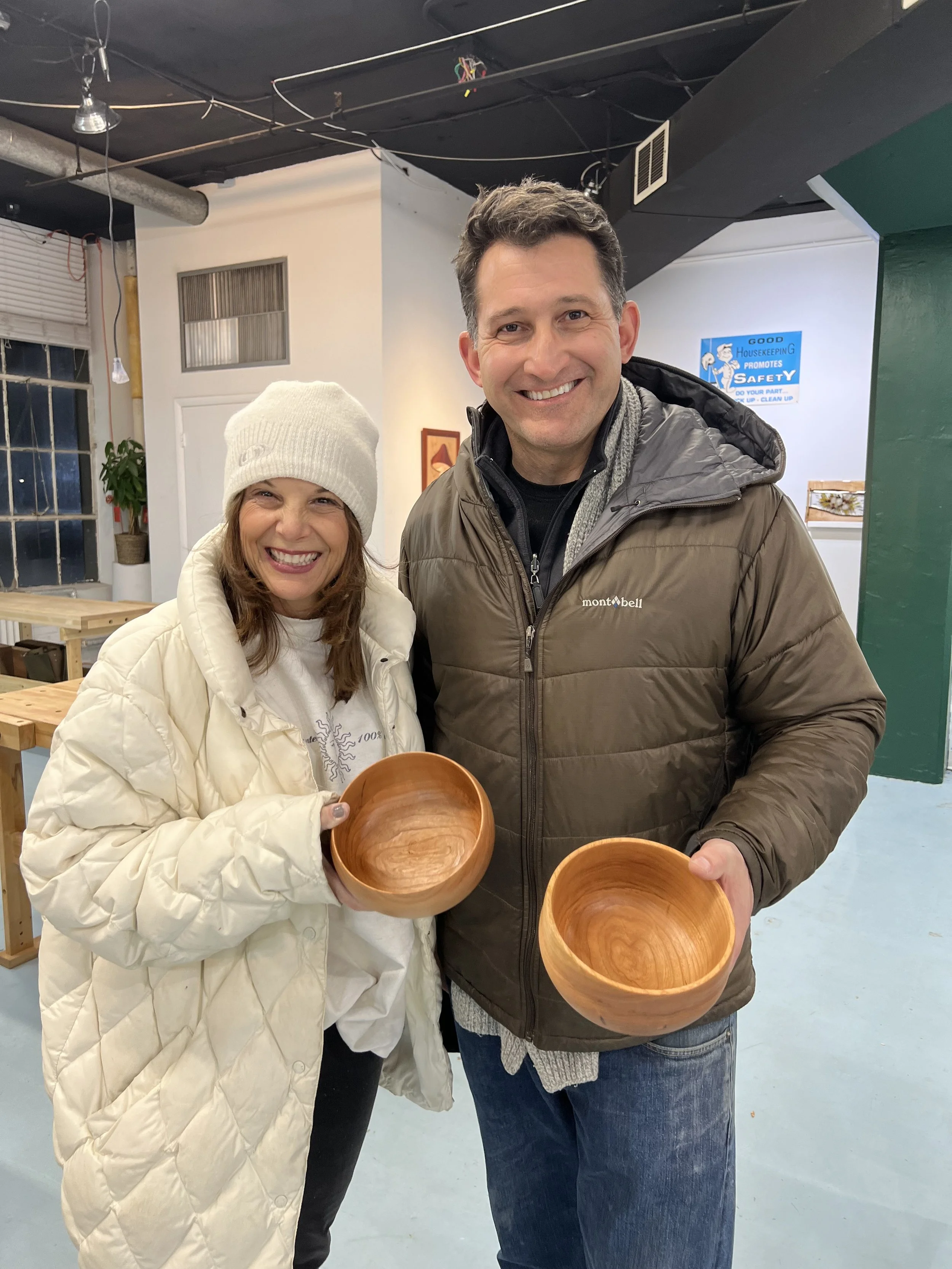 A smiling woman wearing a white beanie and puffy jacket holding a wooden bowl, standing next to a smiling man in a brown jacket also holding a wooden bowl, indoors with a sign about housekeeping and safety in the background.
