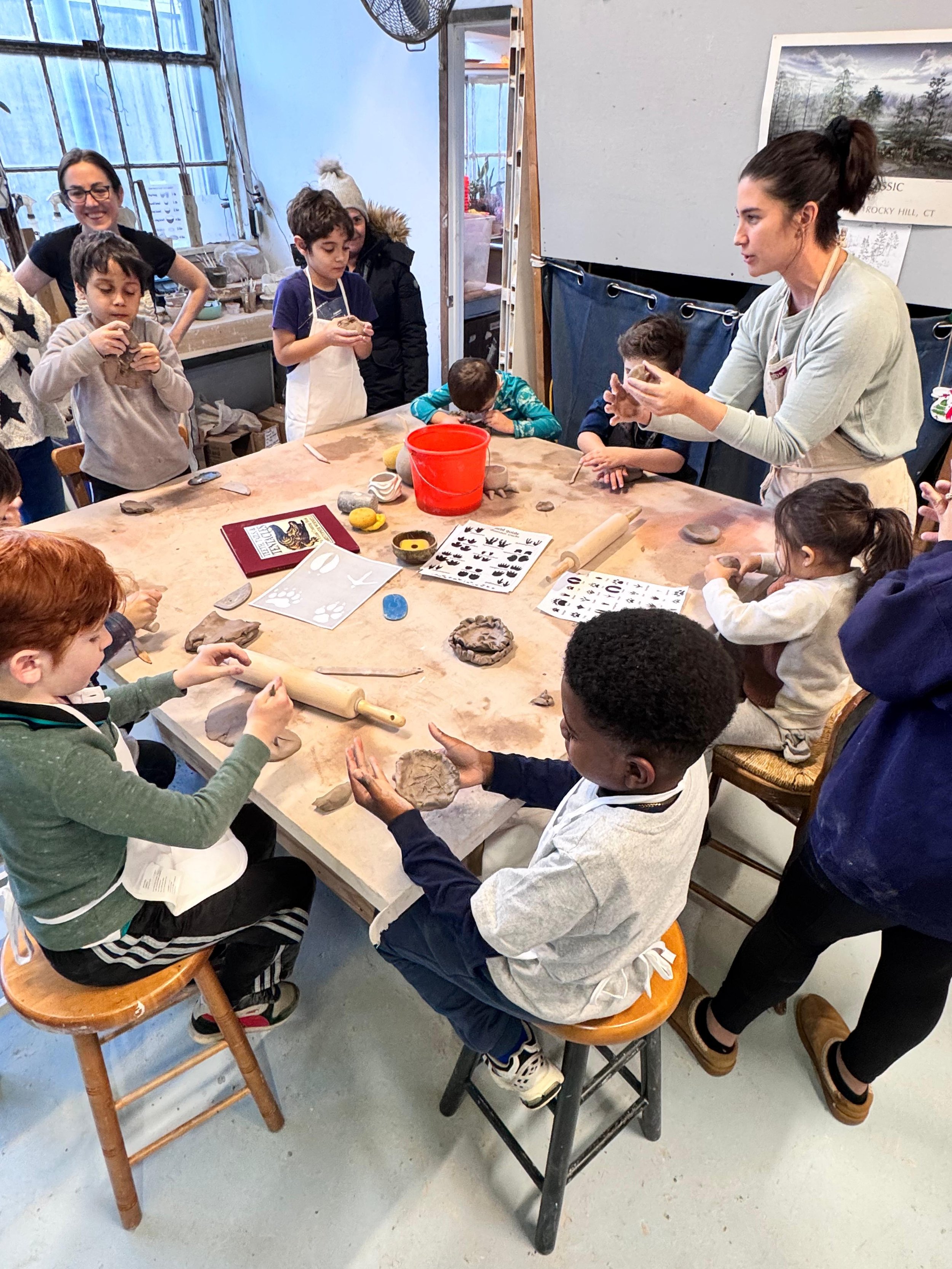 Children and an adult gathered around a table in a pottery class, working with clay and pottery tools.