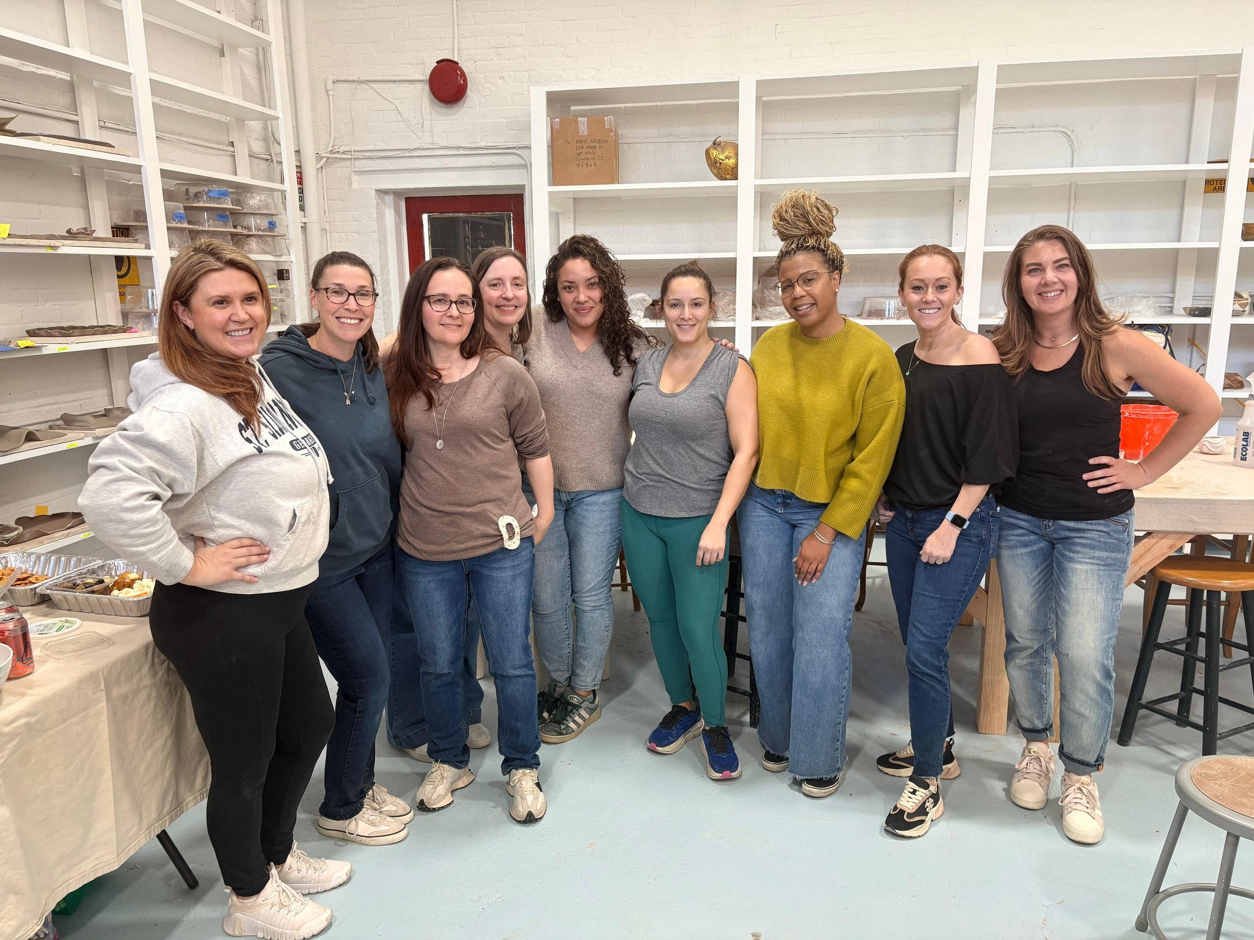 A group of nine women standing together smiling in an indoor space with white brick walls, shelves, and tables with food.