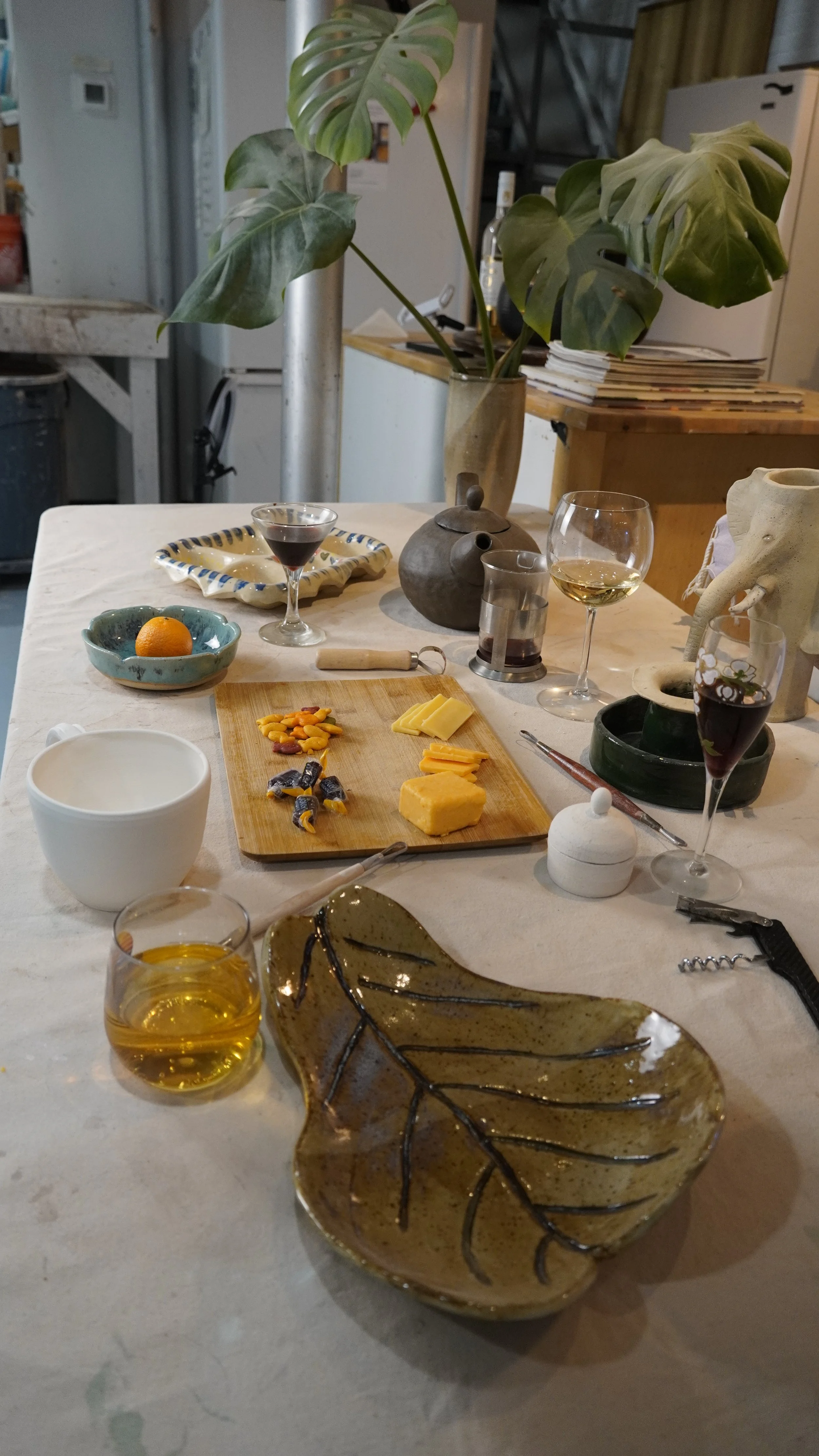 A white table set with cheese, chocolates, a glass of white wine, glasses of red wine, a glass of dark drink, a small white bowl, and decorative plates with a leaf design. In the background, there is a large plant, a bottle of wine, and various kitchen items.