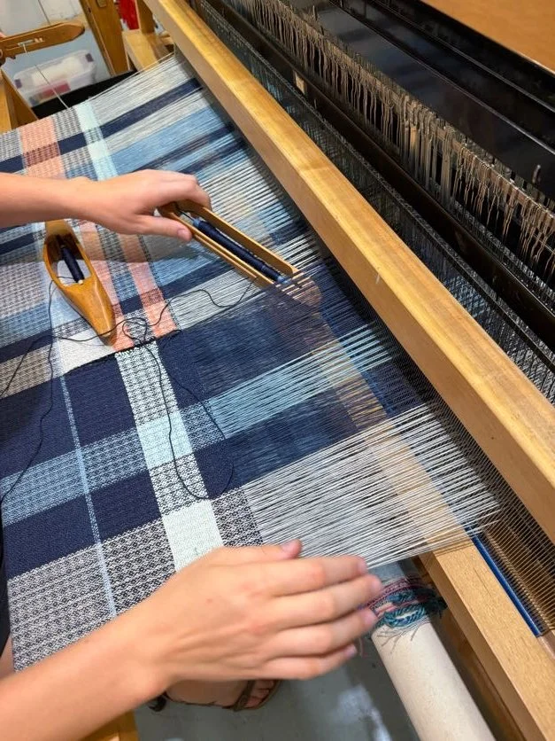 A person weaving a plaid fabric on a loom, using a heddle hook to manage warp threads.