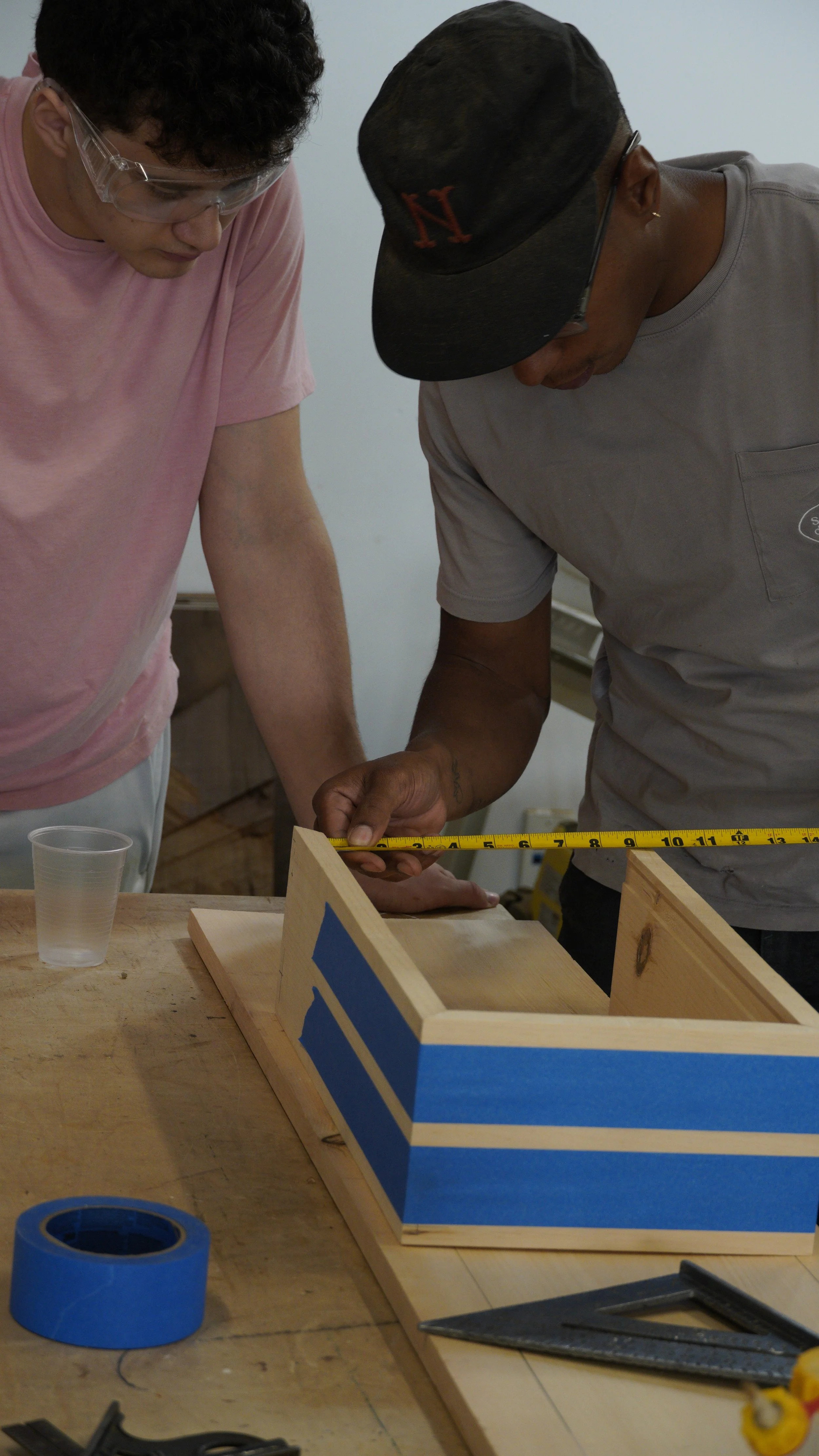 Two men working on a woodworking project at a table, measuring and assembling wooden structures, with tools and blue tape nearby.