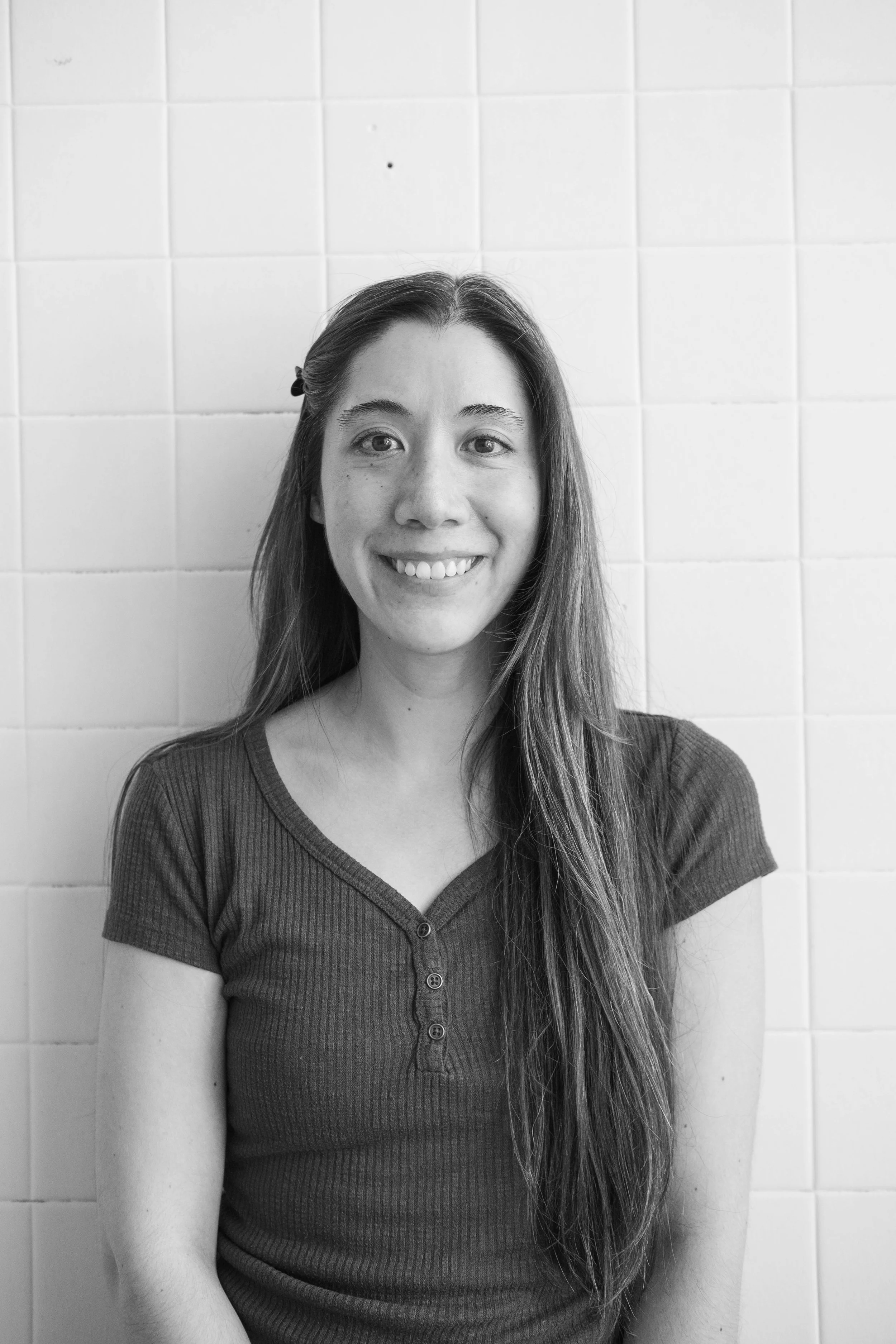 A young woman with long hair and a hair clip, smiling, standing in front of a tiled wall.