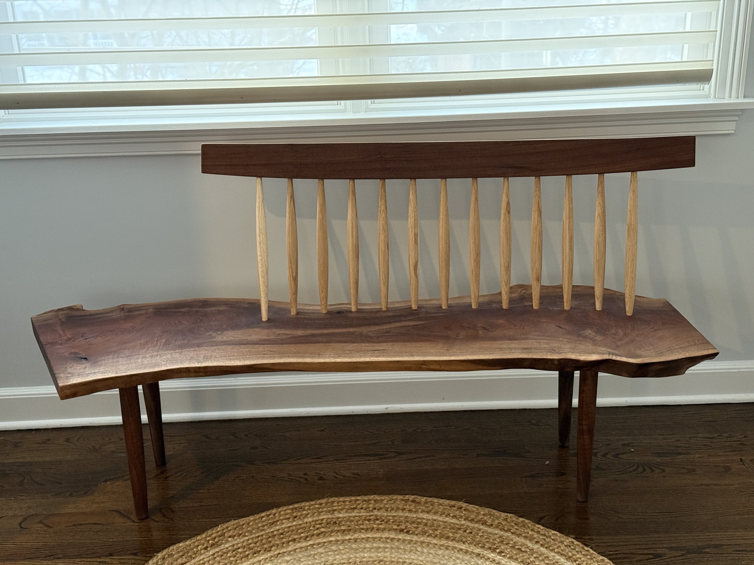 A wooden bench with spindled backrests and a woven rug in front of it, located near a window with white trim.
