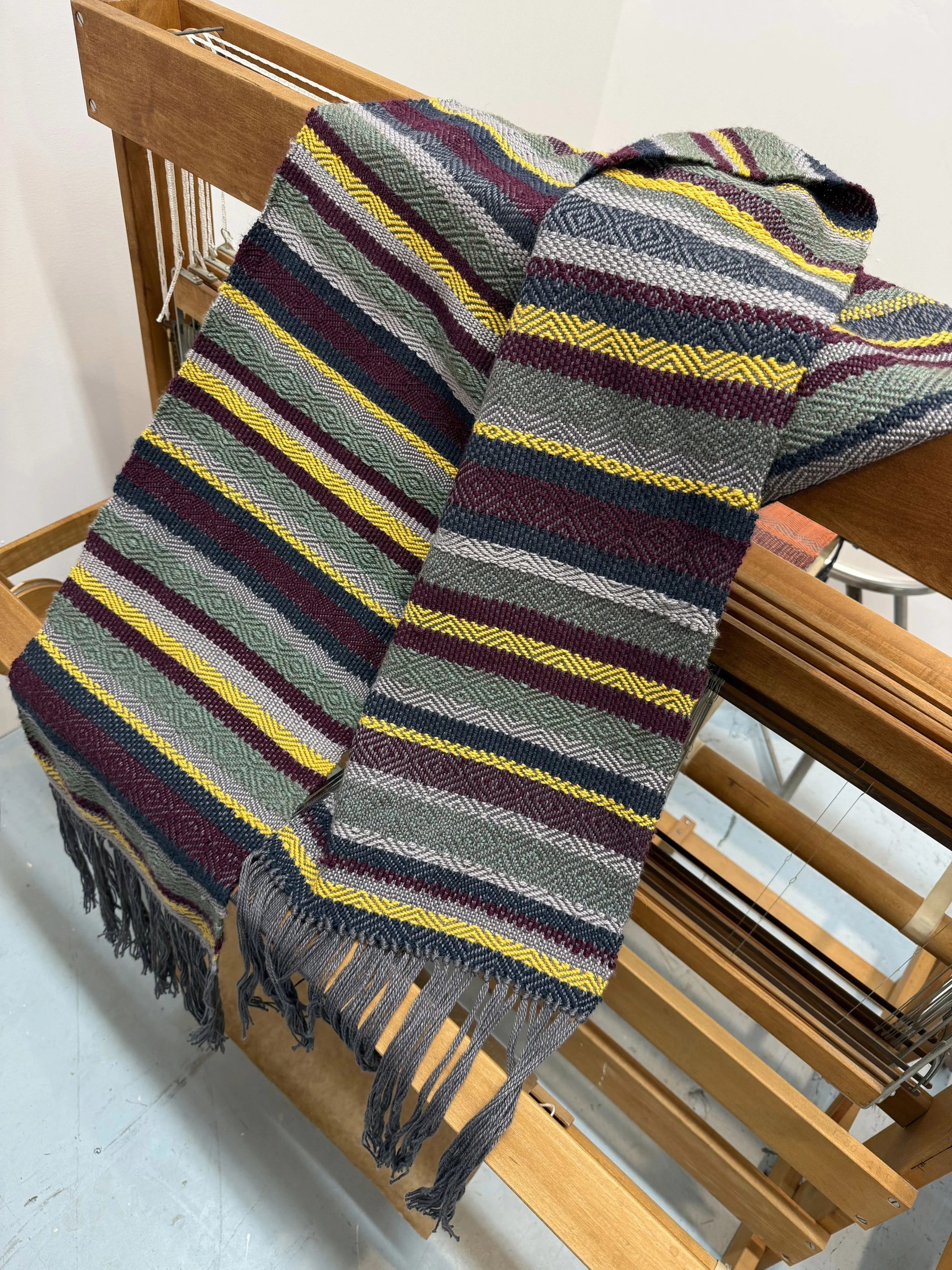 Two woven blankets with colorful striped and geometric patterns, one draped over a wooden weaving loom.
