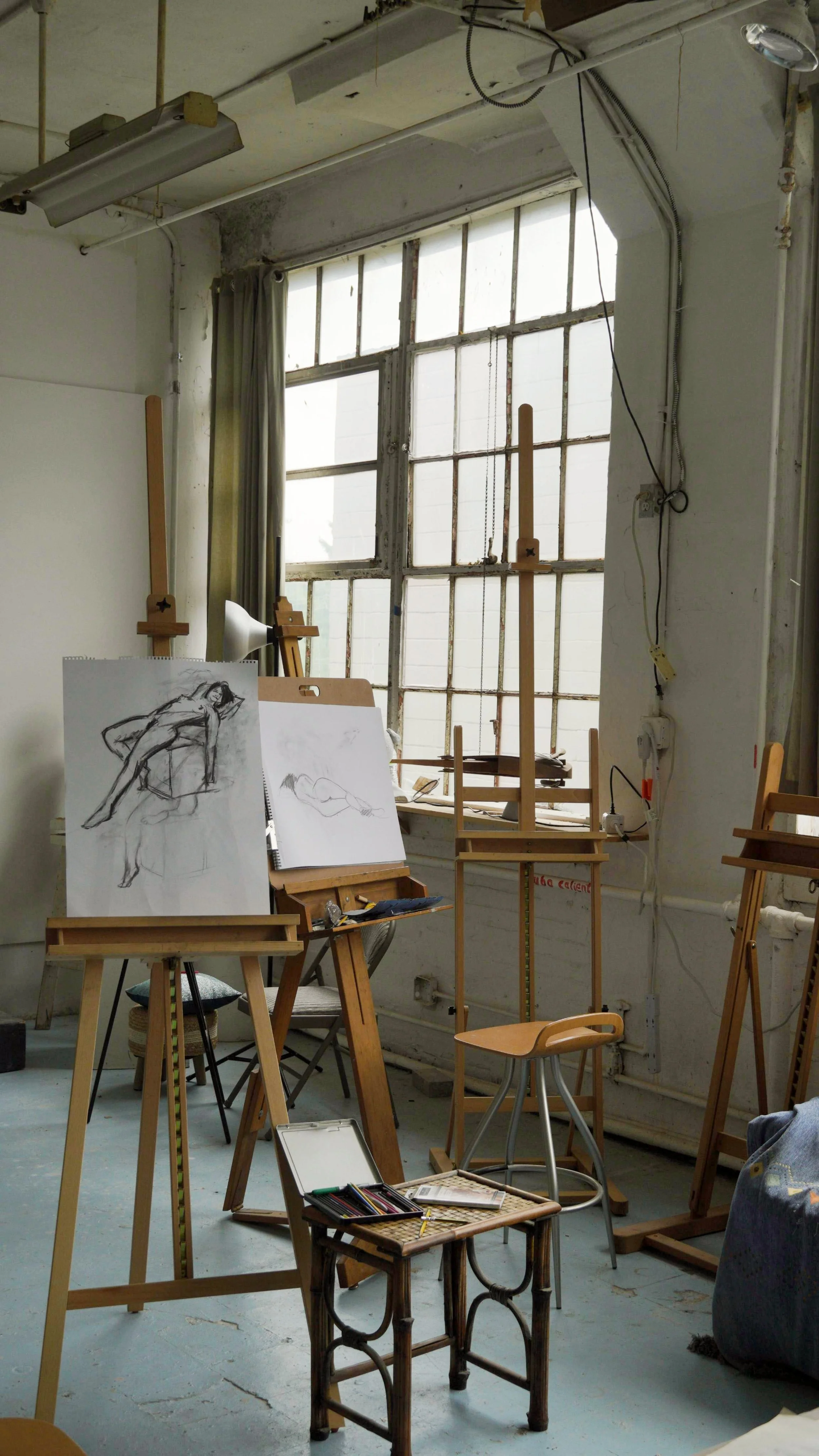 An art studio with easels, sketches, and a large window letting in natural light. The sketches include a reclining female figure drawing.